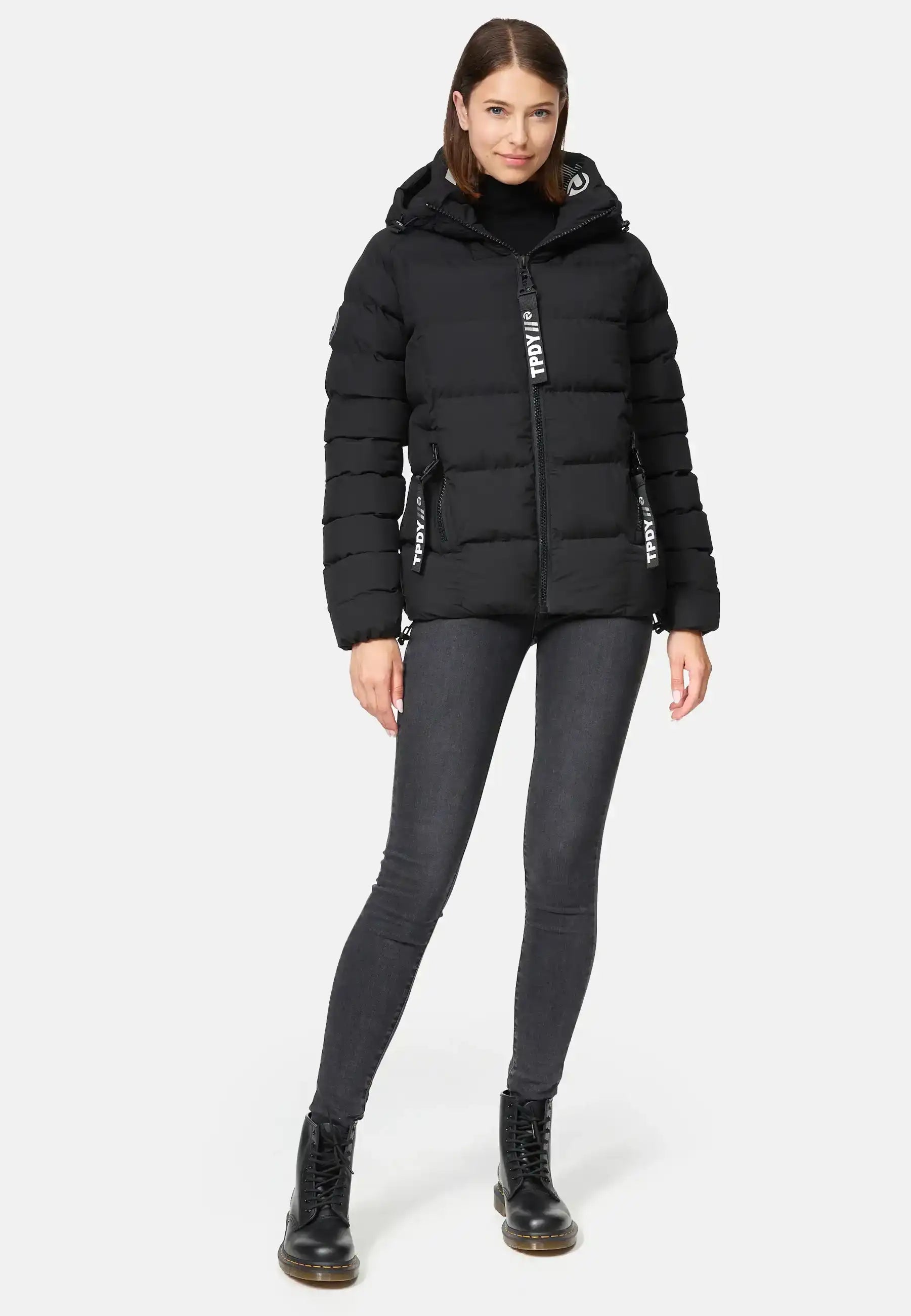 Eine Frau trägt die Murphy F Matte Steppjacke mit Kapuze von trueprodigy, dazu dunkle Skinny Jeans und schwarze Schnürstiefel. Sie steht vor neutralem Hintergrund, blickt nach vorne und setzt den linken Fuß leicht vor den rechten.