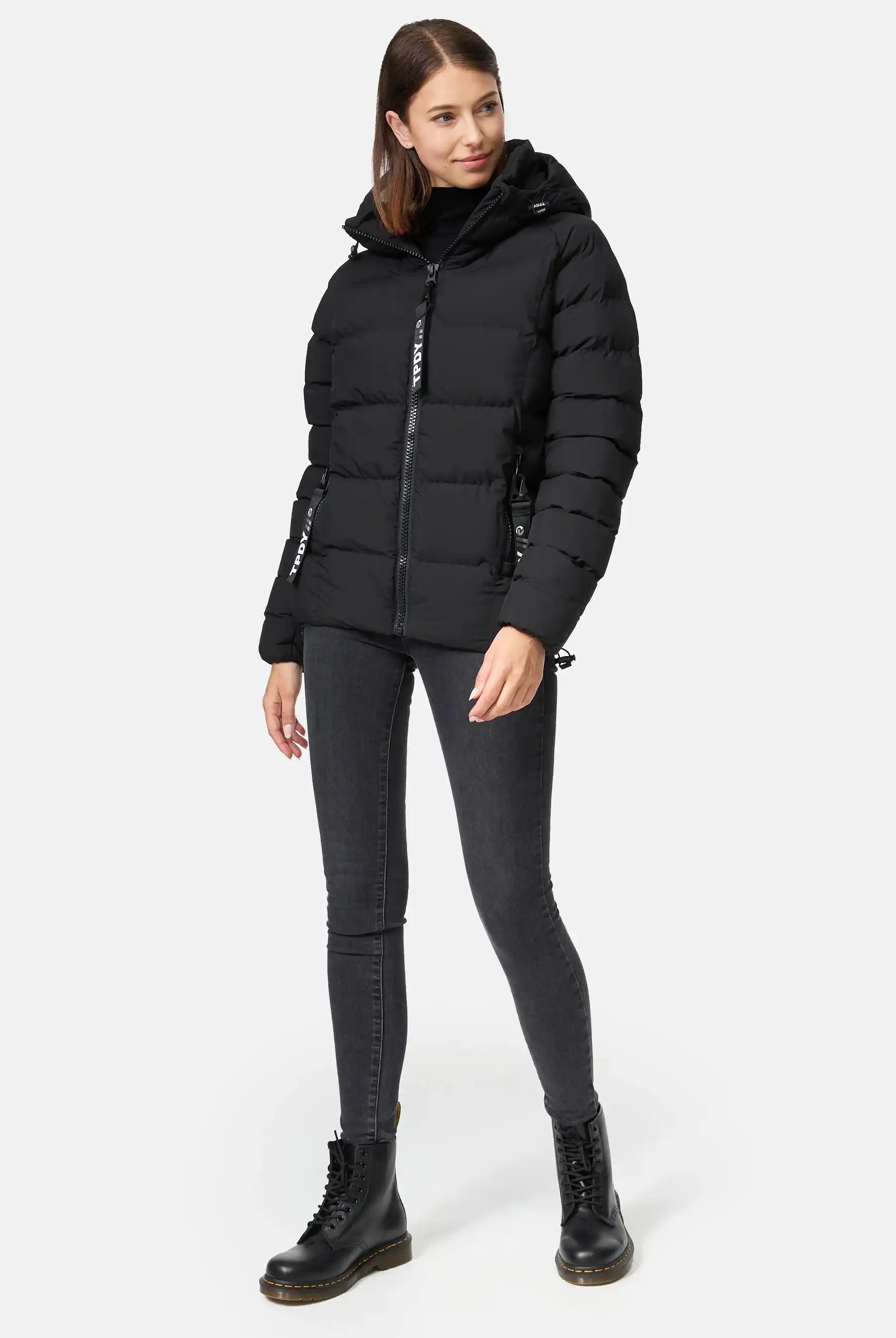 Eine Frau trägt die Murphy F Matte Steppjacke mit Kapuze von trueprodigy, dazu schwarze Skinny Jeans und Schnürstiefel. Sie steht vor neutralem Hintergrund, hat glattes braunes Haar und blickt mit neutralem Ausdruck leicht zur Seite.