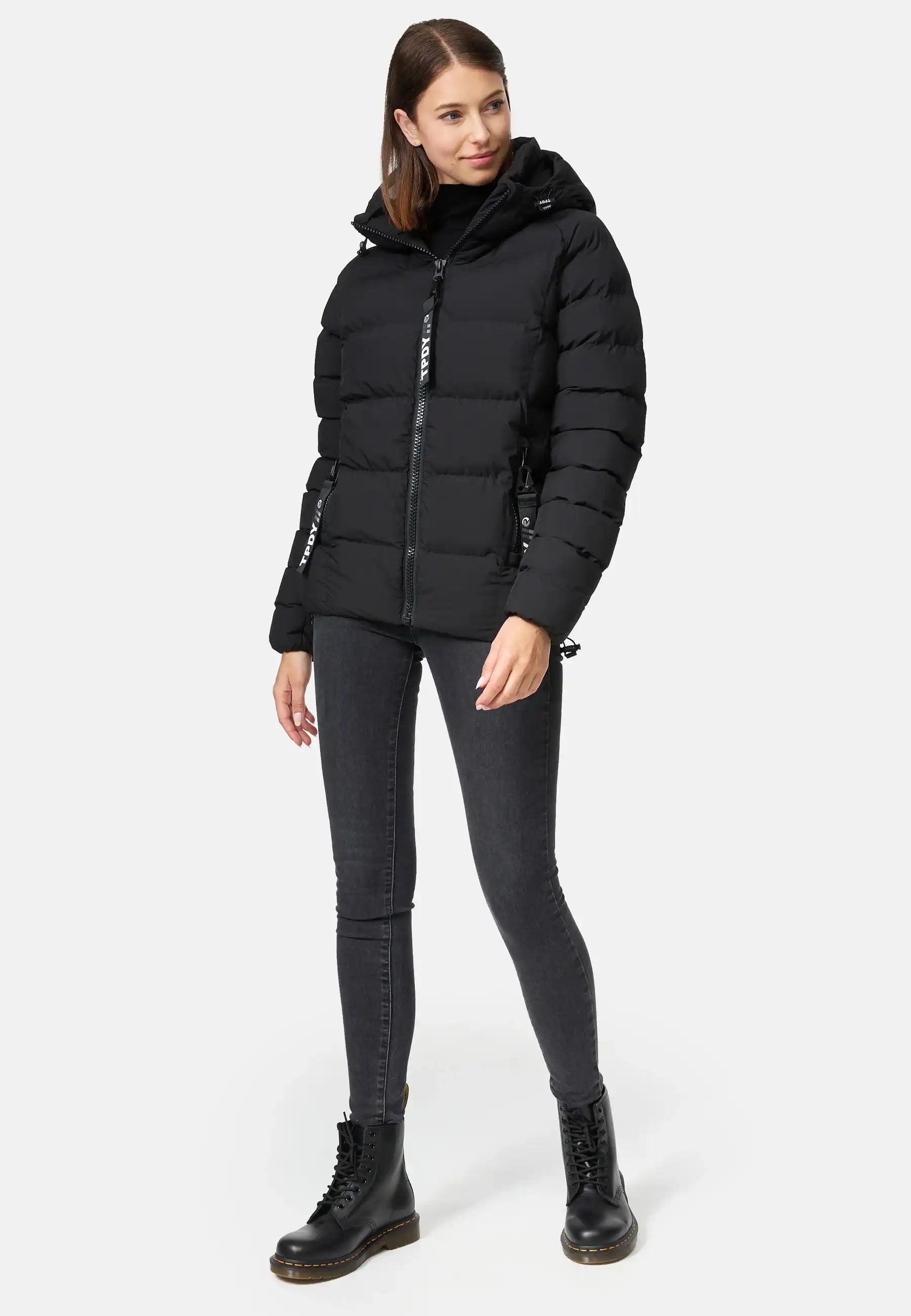 Eine Frau trägt die Murphy F Matte Steppjacke mit Kapuze von trueprodigy, dazu schwarze Skinny Jeans und Schnürstiefel. Sie steht vor neutralem Hintergrund, hat glattes braunes Haar und blickt mit neutralem Ausdruck leicht zur Seite.