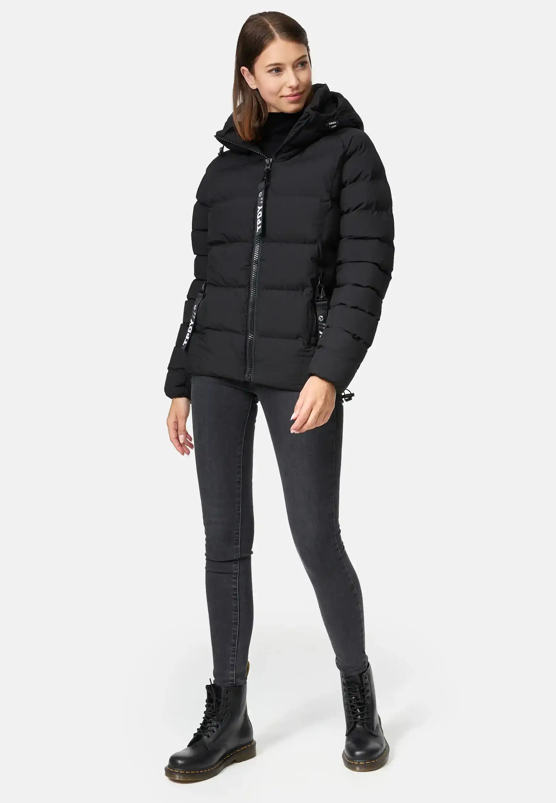 Eine Frau trägt die Murphy F Matte Steppjacke mit Kapuze von trueprodigy, dazu schwarze Skinny Jeans und Schnürstiefel. Sie steht vor neutralem Hintergrund, hat glattes braunes Haar und blickt mit neutralem Ausdruck leicht zur Seite.