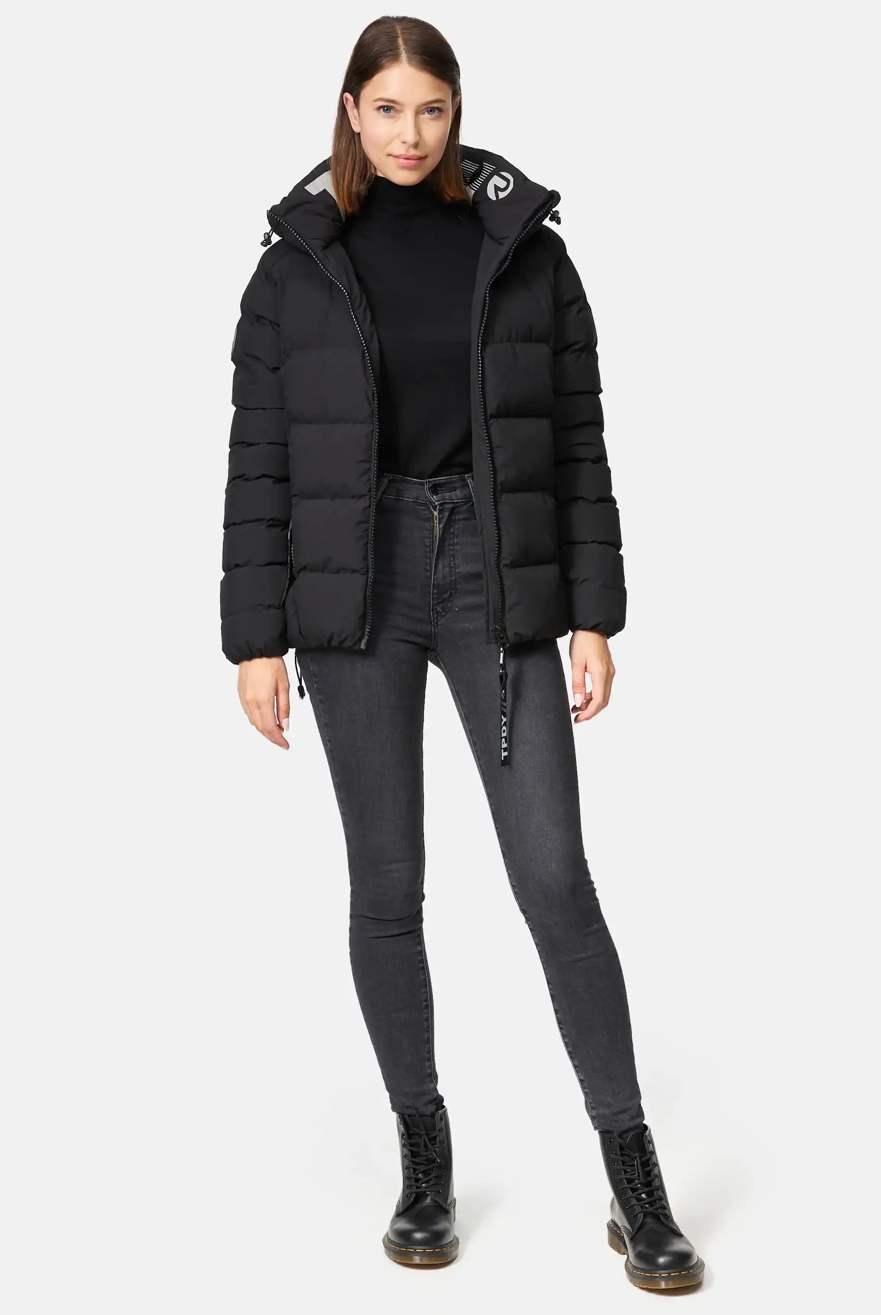 Eine Frau trägt die Murphy F Matte Steppjacke mit Kapuze von trueprodigy, kombiniert mit schwarzem Rollkragen, dunklen Skinny-Jeans und schwarzen Schnürstiefeln. Sie hat glattes, schulterlanges braunes Haar und einen neutralen Ausdruck.