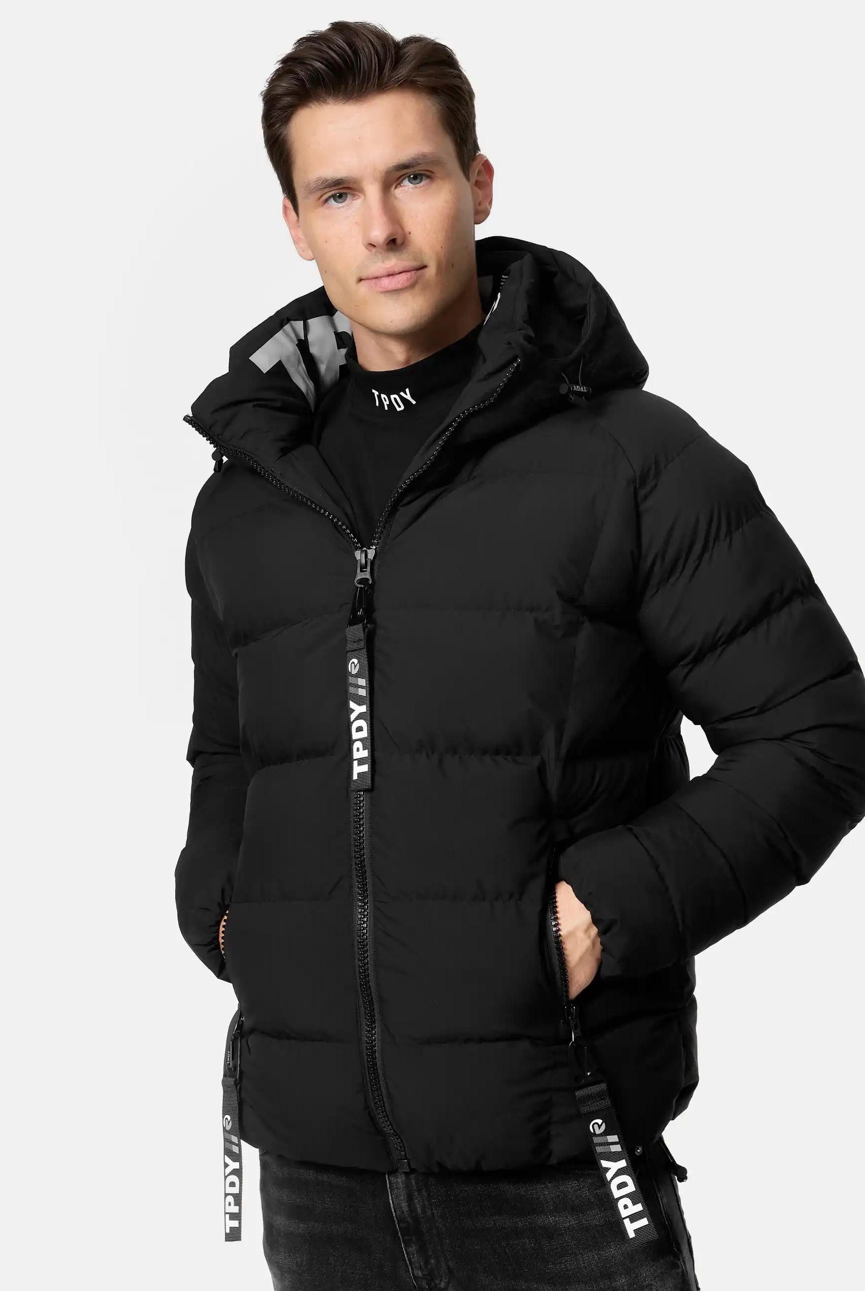 Eine Person trägt die Murphy Matte Steppjacke mit Kapuze von trueprodigy in Schwarz, steht frontal vor weißem Hintergrund; beide Hände sind in den Taschen, die wasserabweisende Jacke zeigt sichtbare Reißverschlüsse.