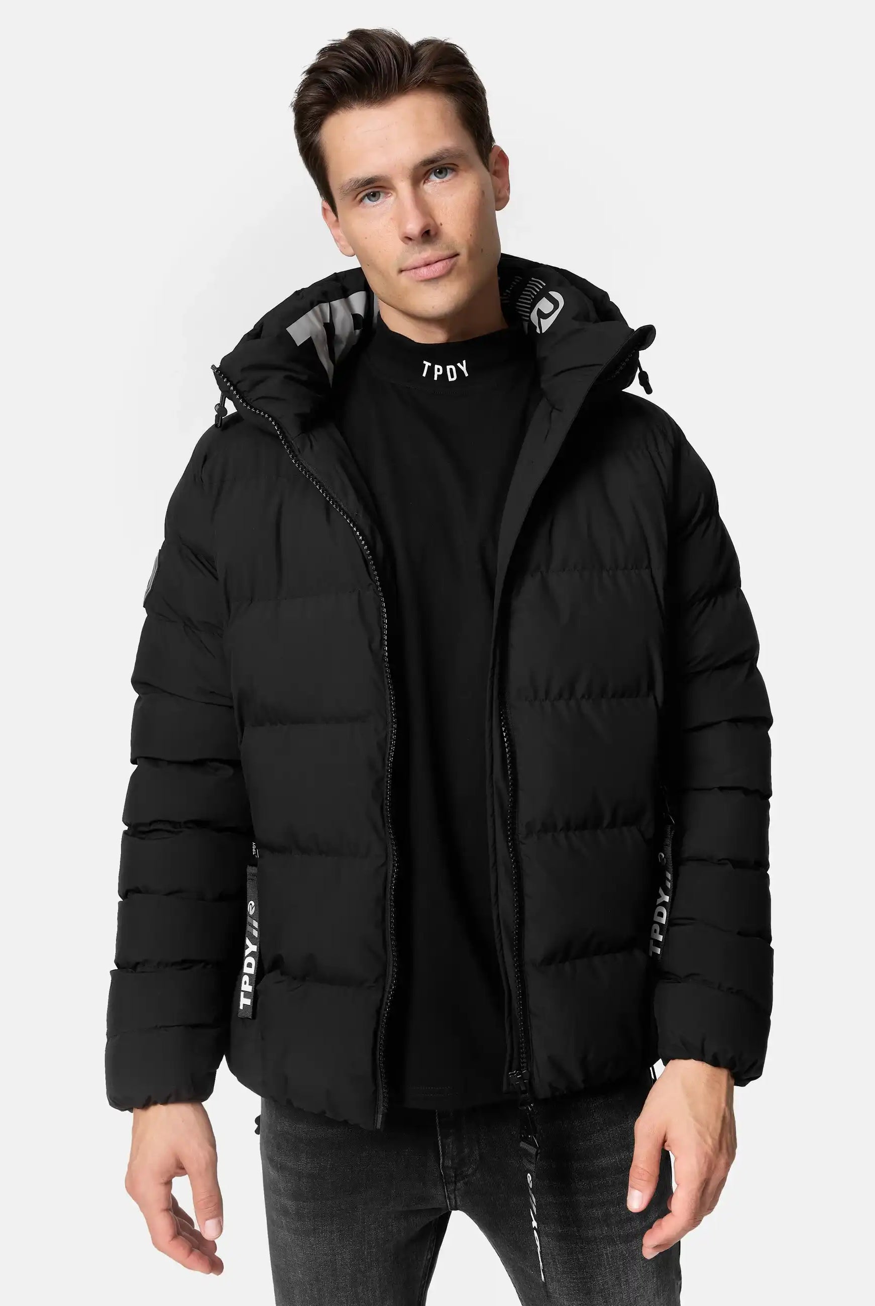 Ein Mann, der eine Trueprodigy Murphy Matte Steppjacke mit Kapuze über einem schwarzen T-Shirt und einer dunklen Jeans trägt, steht der Kamera vor einem schlichten weißen Hintergrund gegenüber.