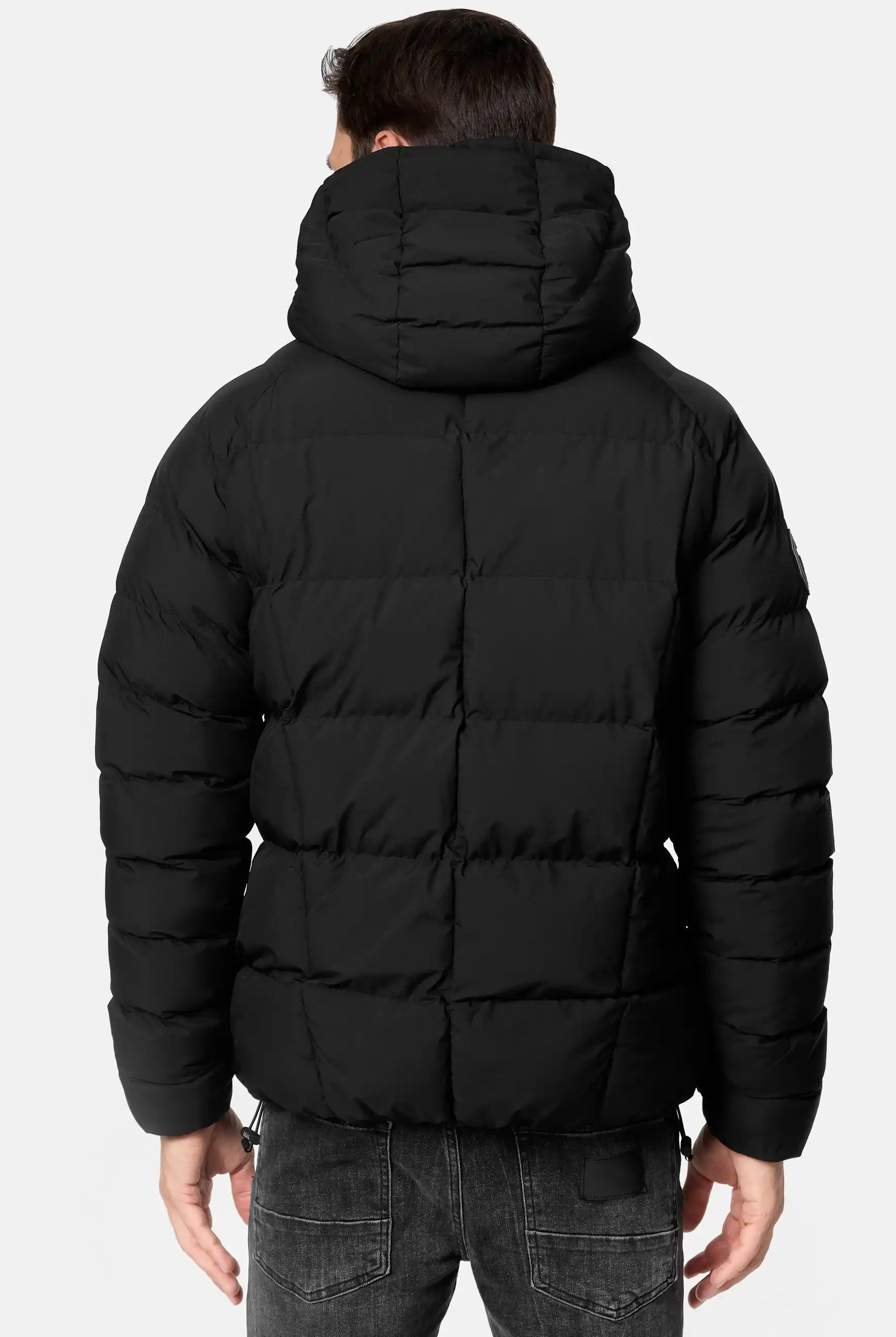 Eine Person ist von hinten in einer Murphy Matte Steppjacke mit Kapuze von trueprodigy zu sehen. Die wasserabweisende Jacke ist horizontal gesteppt und wird mit dunkelgrauen Jeans vor einem hellen Hintergrund kombiniert.