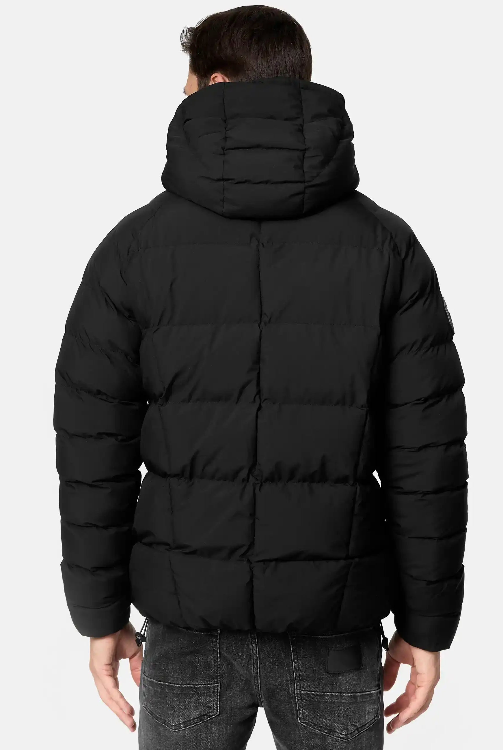 Eine Person ist von hinten in einer Murphy Matte Steppjacke mit Kapuze von trueprodigy zu sehen. Die wasserabweisende Jacke ist horizontal gesteppt und wird mit dunkelgrauen Jeans vor einem hellen Hintergrund kombiniert.