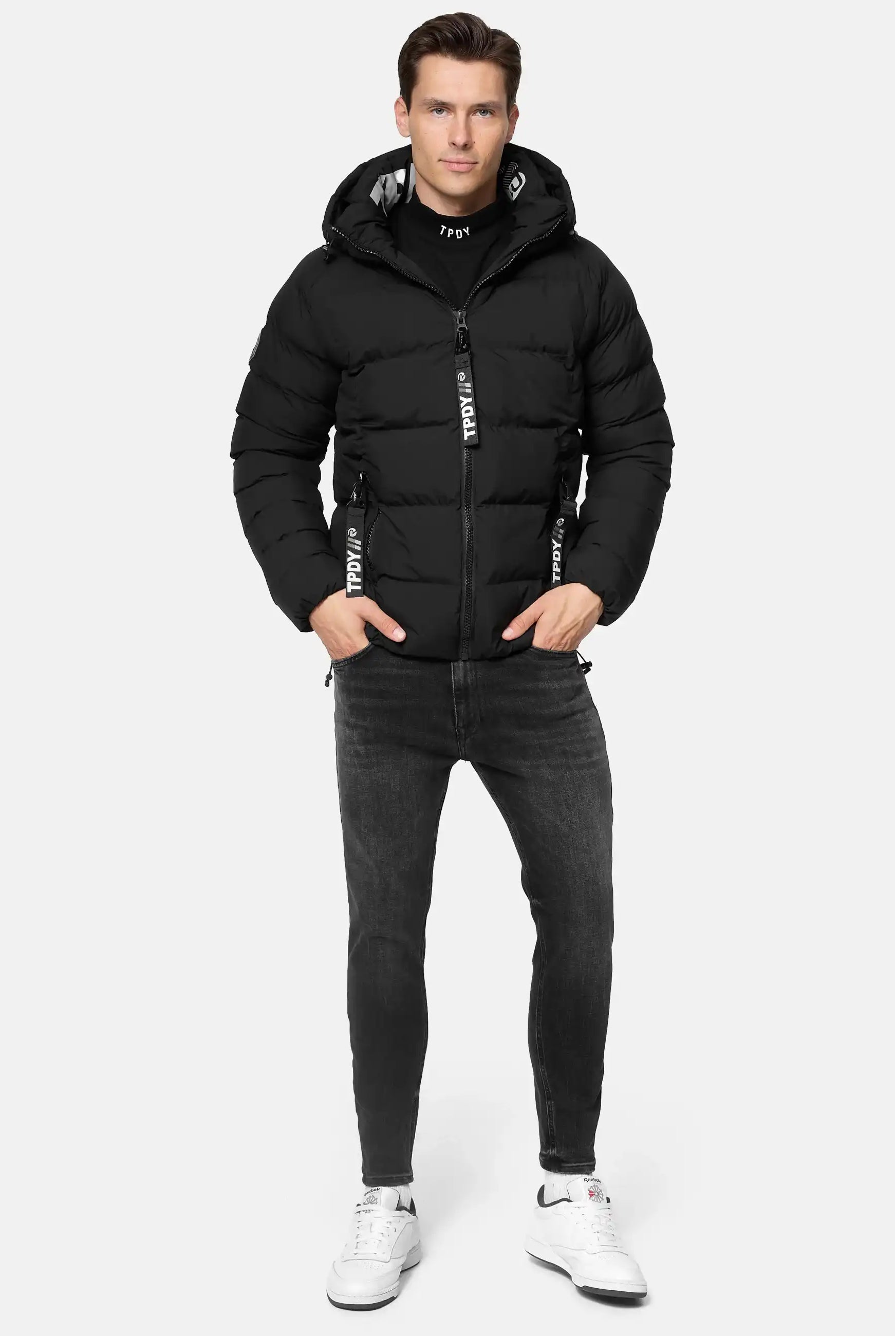 Ein Mann trägt die Murphy Matte Steppjacke mit Kapuze von trueprodigy, schwarze Jeans und weiße Sneakers. Mit den Händen in den Jackentaschen steht er vor einem schlichten, hellgrauen Hintergrund.