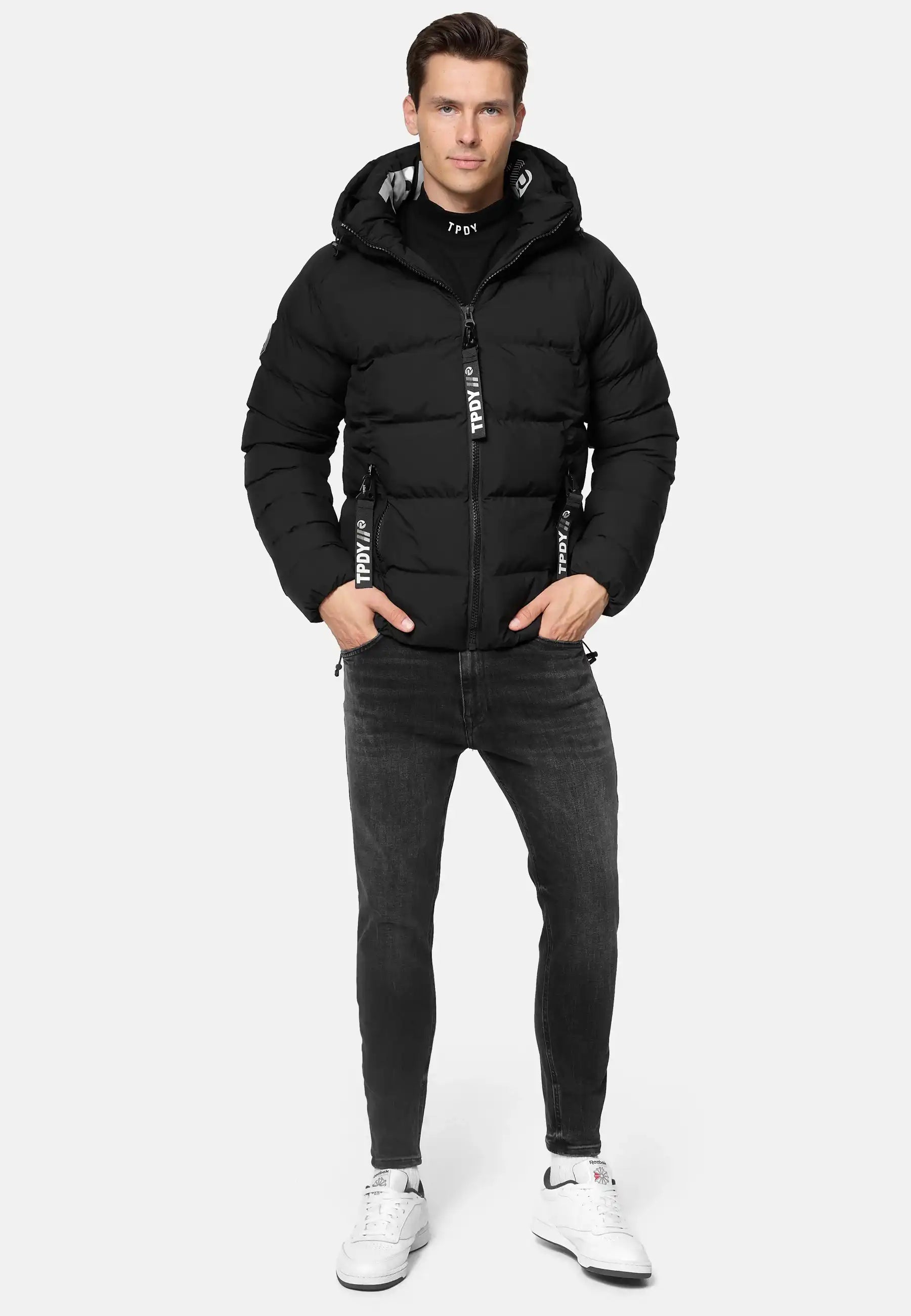 Ein Mann trägt die Murphy Matte Steppjacke mit Kapuze von trueprodigy, schwarze Jeans und weiße Sneakers. Mit den Händen in den Jackentaschen steht er vor einem schlichten, hellgrauen Hintergrund.