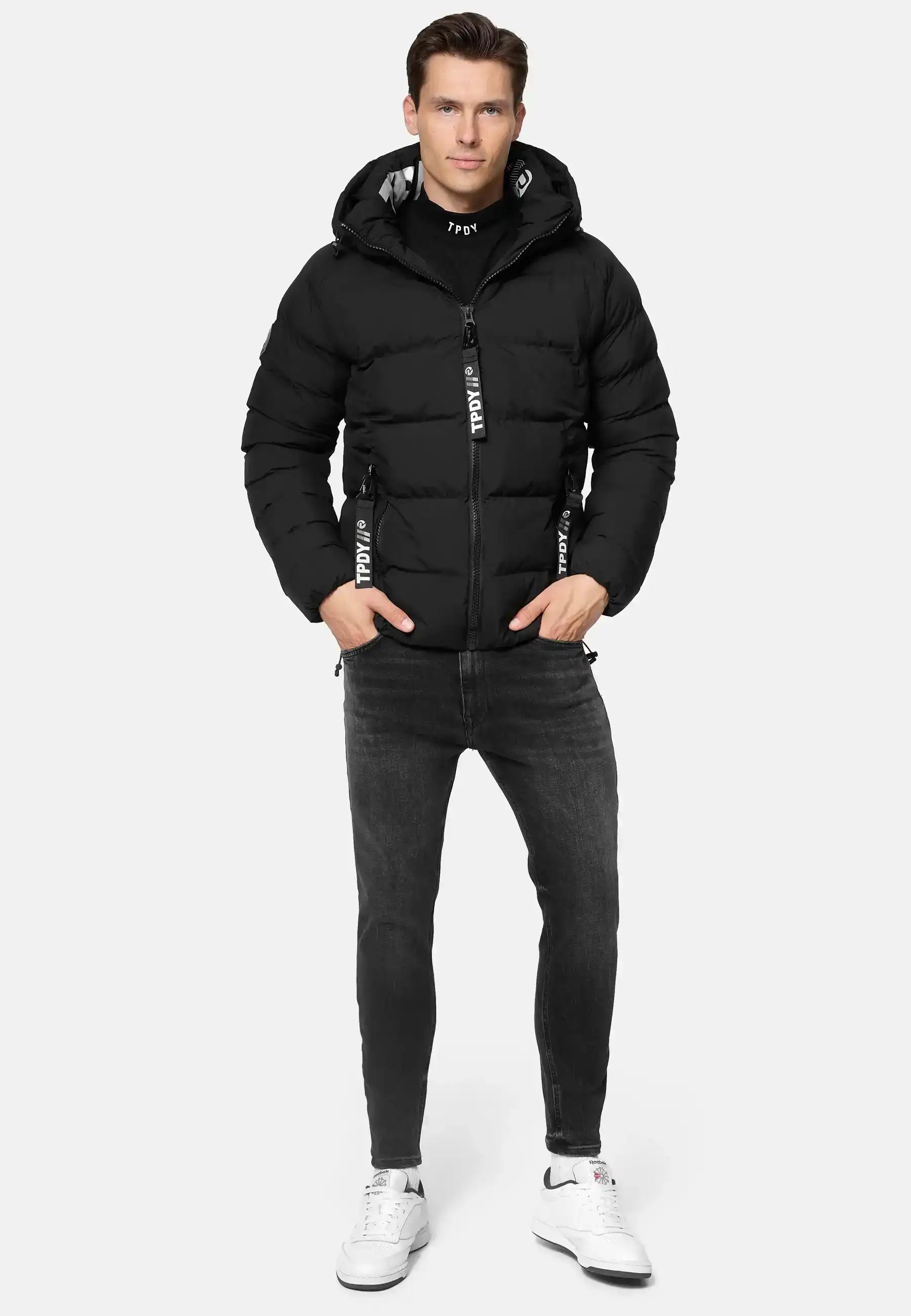 Ein Mann trägt die Murphy Matte Steppjacke mit Kapuze von trueprodigy, schwarze Jeans und weiße Sneakers. Mit den Händen in den Jackentaschen steht er vor einem schlichten, hellgrauen Hintergrund.