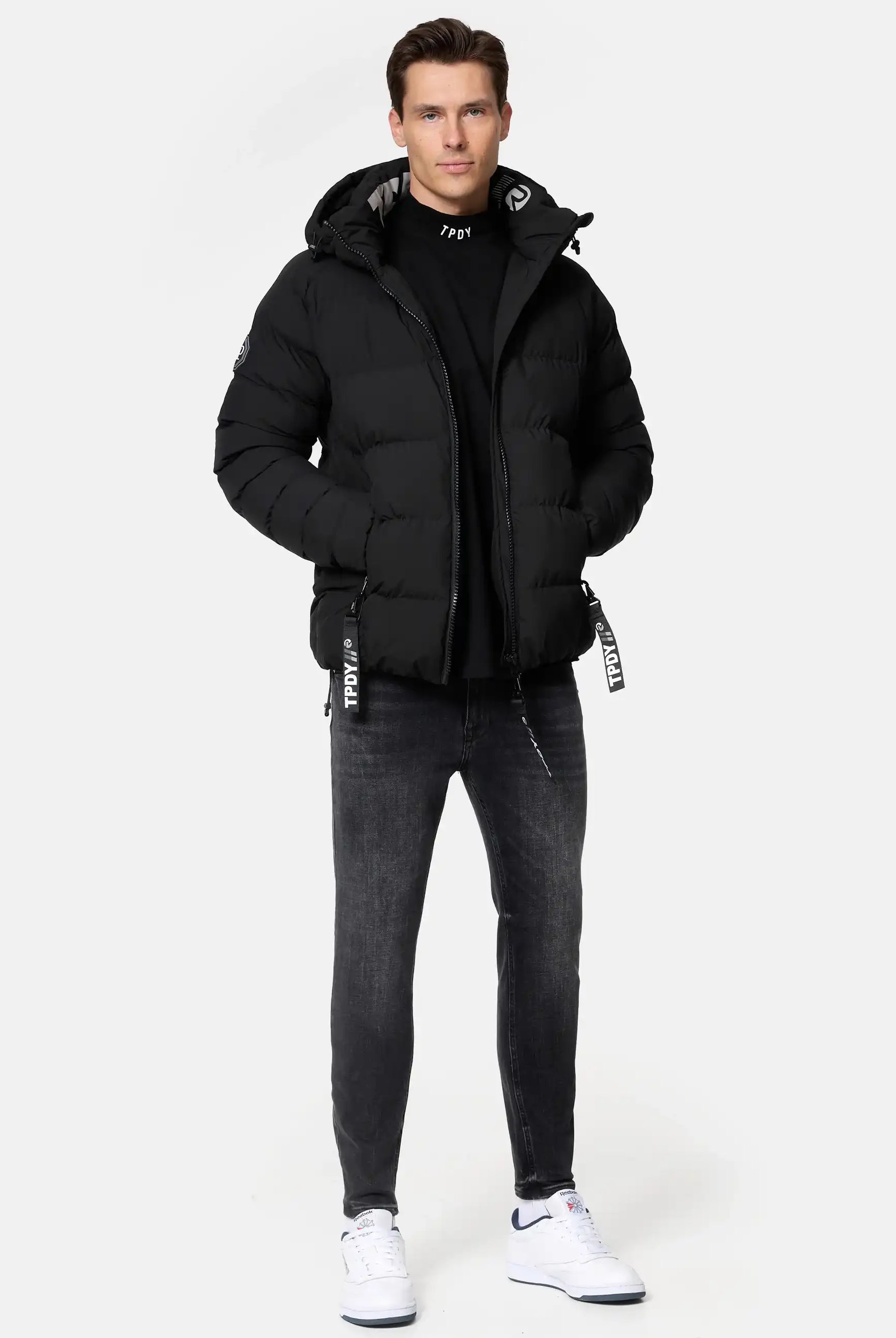 Ein Mann trägt die Murphy Matte Steppjacke mit Kapuze von trueprodigy, ein schwarzes Shirt, dunkelgraue Jeans und weiße Sneakers mit schwarzen Akzenten. Er hat kurze braune Haare und die Hände in den Jackentaschen.