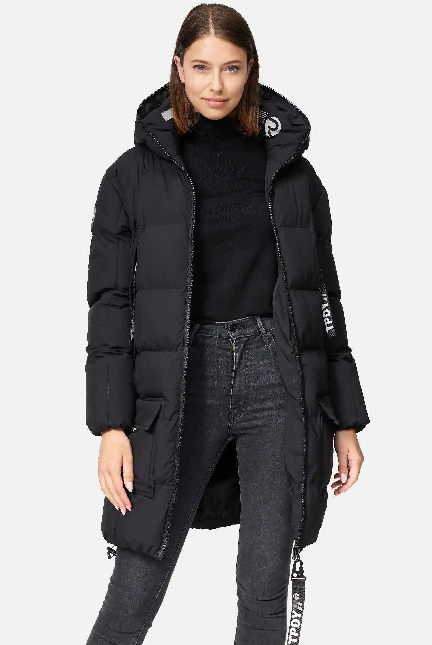 Eine Frau mit glattem braunem Haar blickt nach vorne und trägt die trueprodigy Willo F Lange Pufferjacke mit Kapuze, einen langen schwarzen Mantel mit Reißverschlüssen, Taschen, weißen Textdetails am Ärmel und Träger, über einem schwarzen Rollkragenpullover und dunklen Jeans.