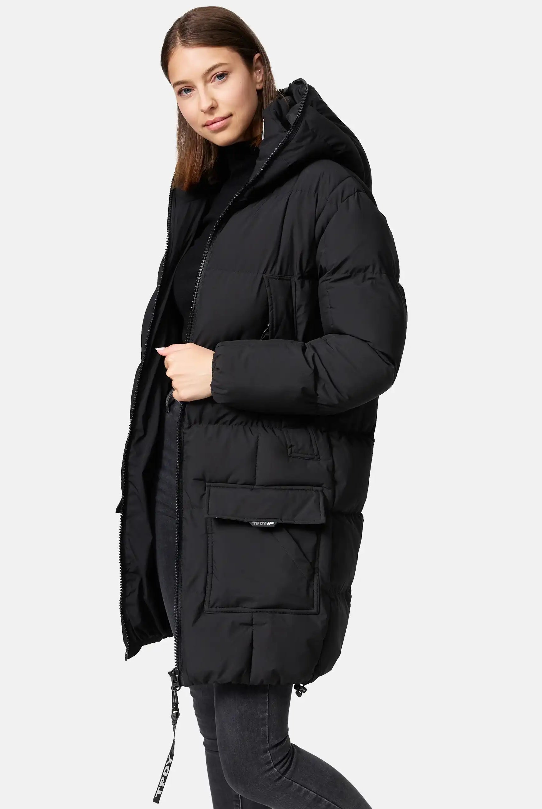 Eine Frau mit glattem braunem Haar trägt die trueprodigy Willo F Lange Pufferjacke mit Kapuze in Schwarz über einer dunklen Jeans, wobei sie leicht zur Seite schaut und den Reißverschluss festhält. Der Hintergrund ist einfarbig hellgrau.