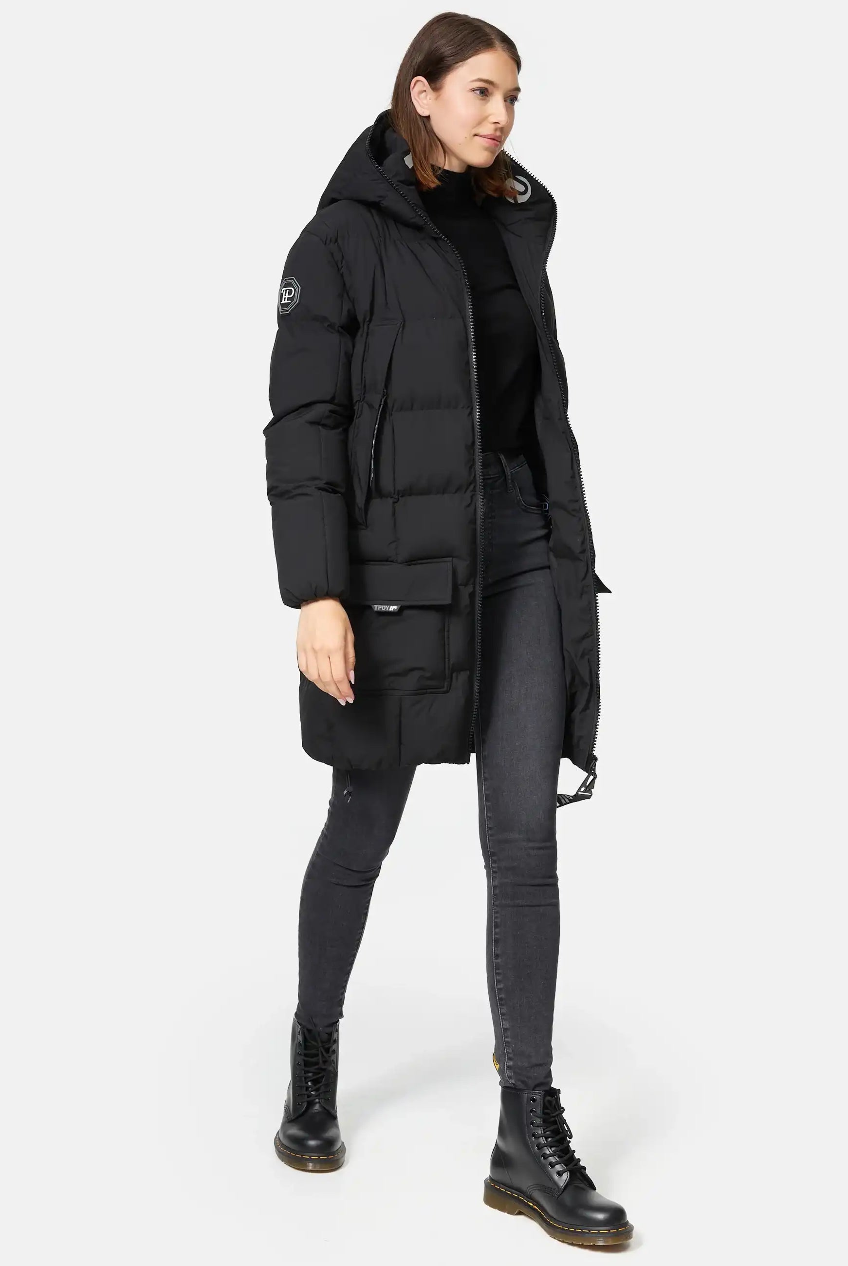 Eine Person, die die trueprodigy Willo F Lange Pufferjacke mit Kapuze in Schwarz trägt, gestylt mit einem schwarzen Rollkragenpullover, dunklen Skinny Jeans und schwarzen Schnürstiefeln, steht vor einem schlichten weißen Hintergrund.