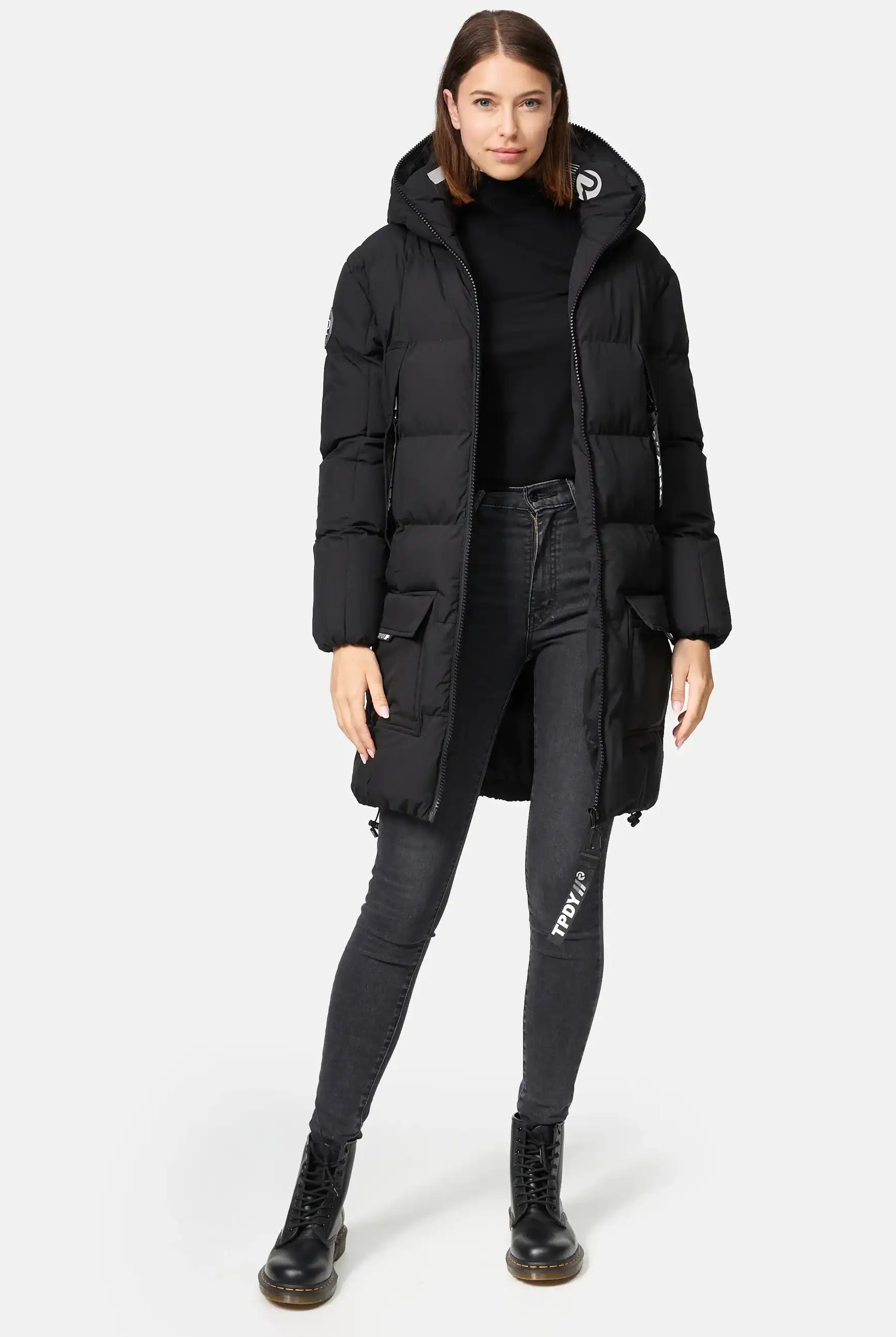 Eine Frau steht vor einem schlichten Hintergrund und trägt die trueprodigy Willo F Lange Pufferjacke mit Kapuze, gepaart mit einem schwarzen Rollkragenpullover, dunklen Skinny Jeans und schwarzen Schnürstiefeln. Ihre Hände liegen entspannt an ihren Seiten.