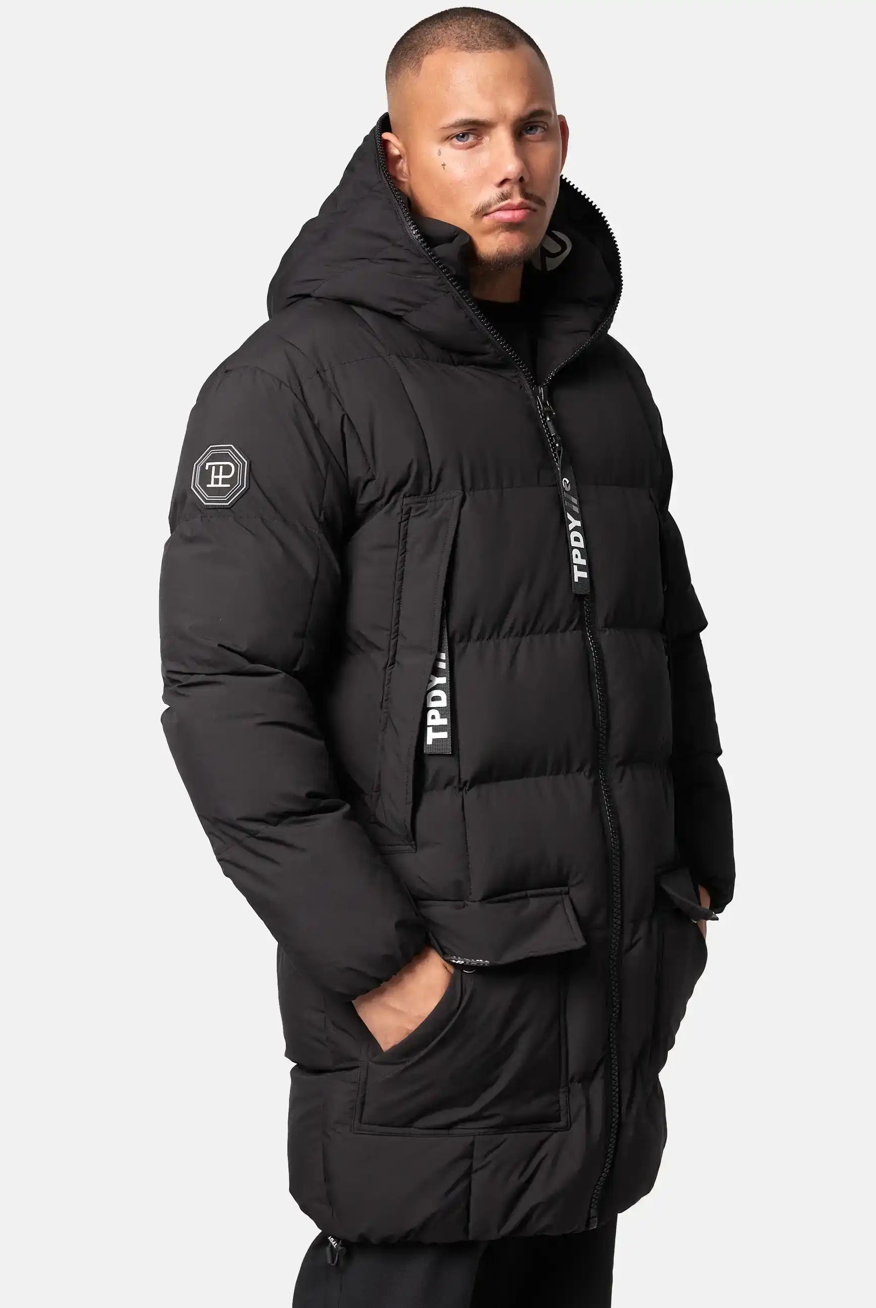 Eine Person trägt die trueprodigy Willo Lange Pufferjacke mit Kapuze, eine lange schwarze Herrenjacke mit einem Logoaufnäher am Ärmel und Marken-Reißverschlüssen. Eine Hand steckt in einer Tasche. Der Hintergrund ist einfarbig und hell.