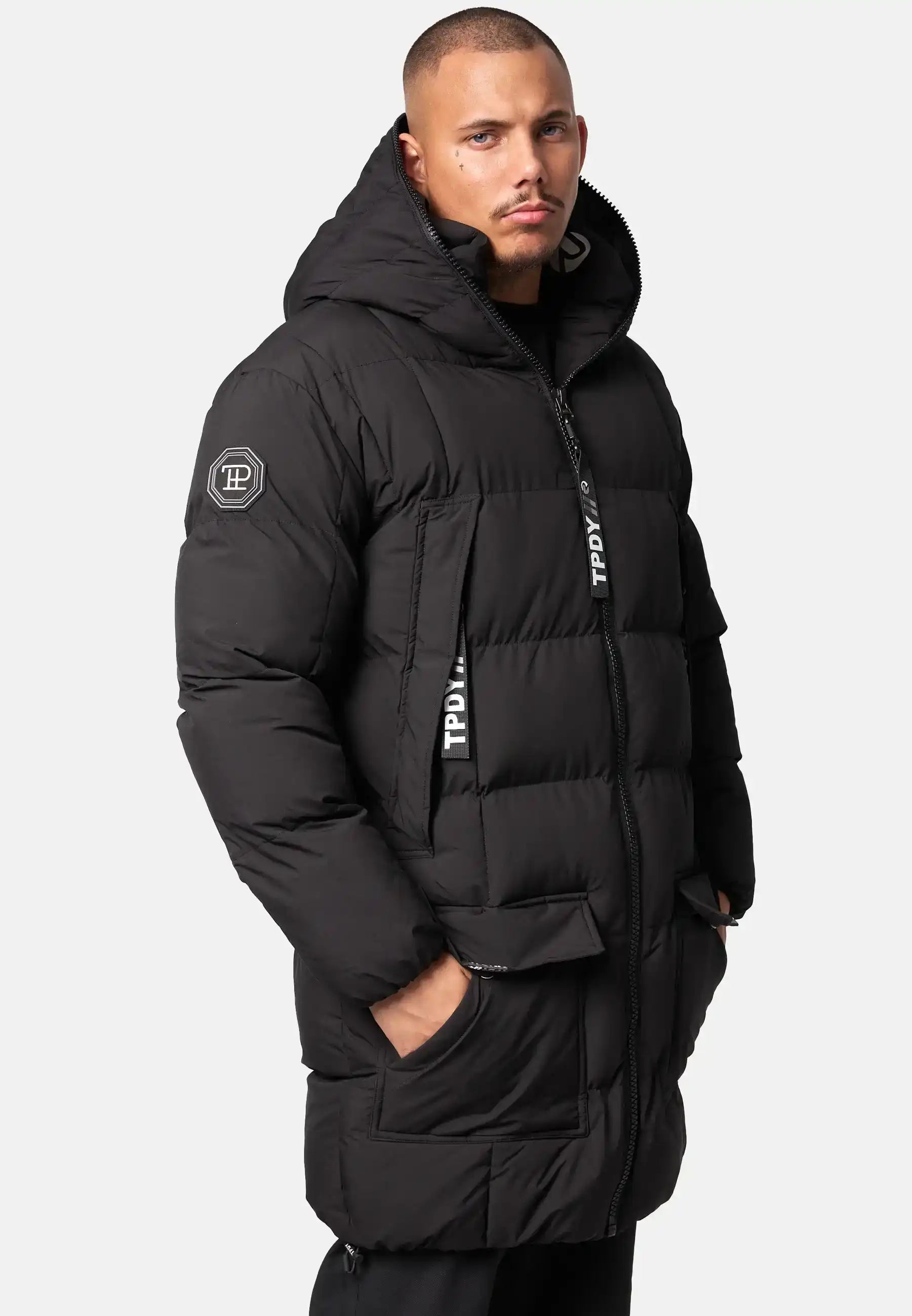 Eine Person trägt die trueprodigy Willo Lange Pufferjacke mit Kapuze, eine lange schwarze Herrenjacke mit einem Logoaufnäher am Ärmel und Marken-Reißverschlüssen. Eine Hand steckt in einer Tasche. Der Hintergrund ist einfarbig und hell.