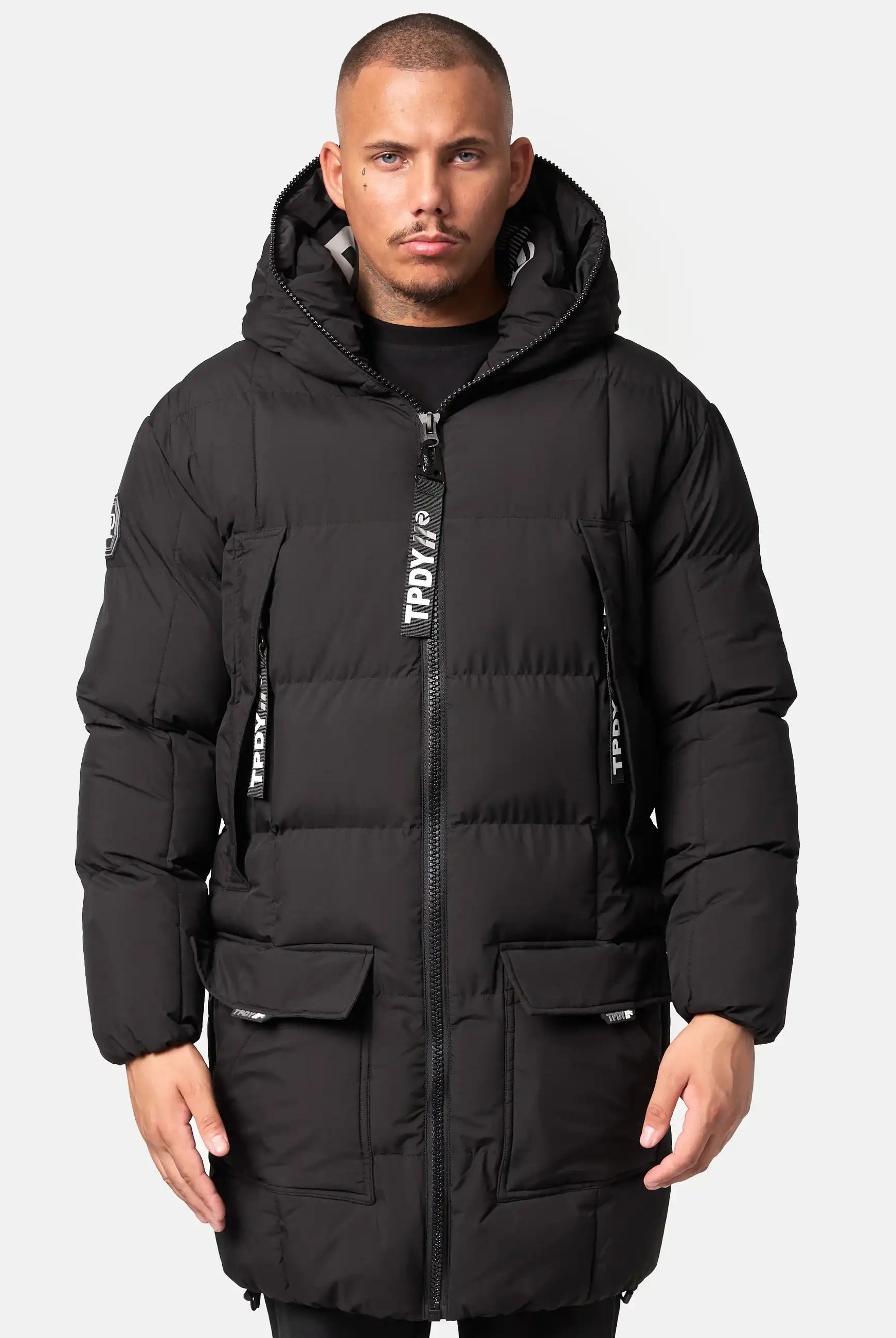 Ein Mann blickt nach vorne und trägt die schwarze Trueprodigy Willo Lange Pufferjacke mit Kapuze. Sie hat eine Kapuze, einen Frontreißverschluss, zwei Brusttaschen, zwei große Pattentaschen und weiße Logodetails an den Reißverschlusszügen und am linken Ärmel.