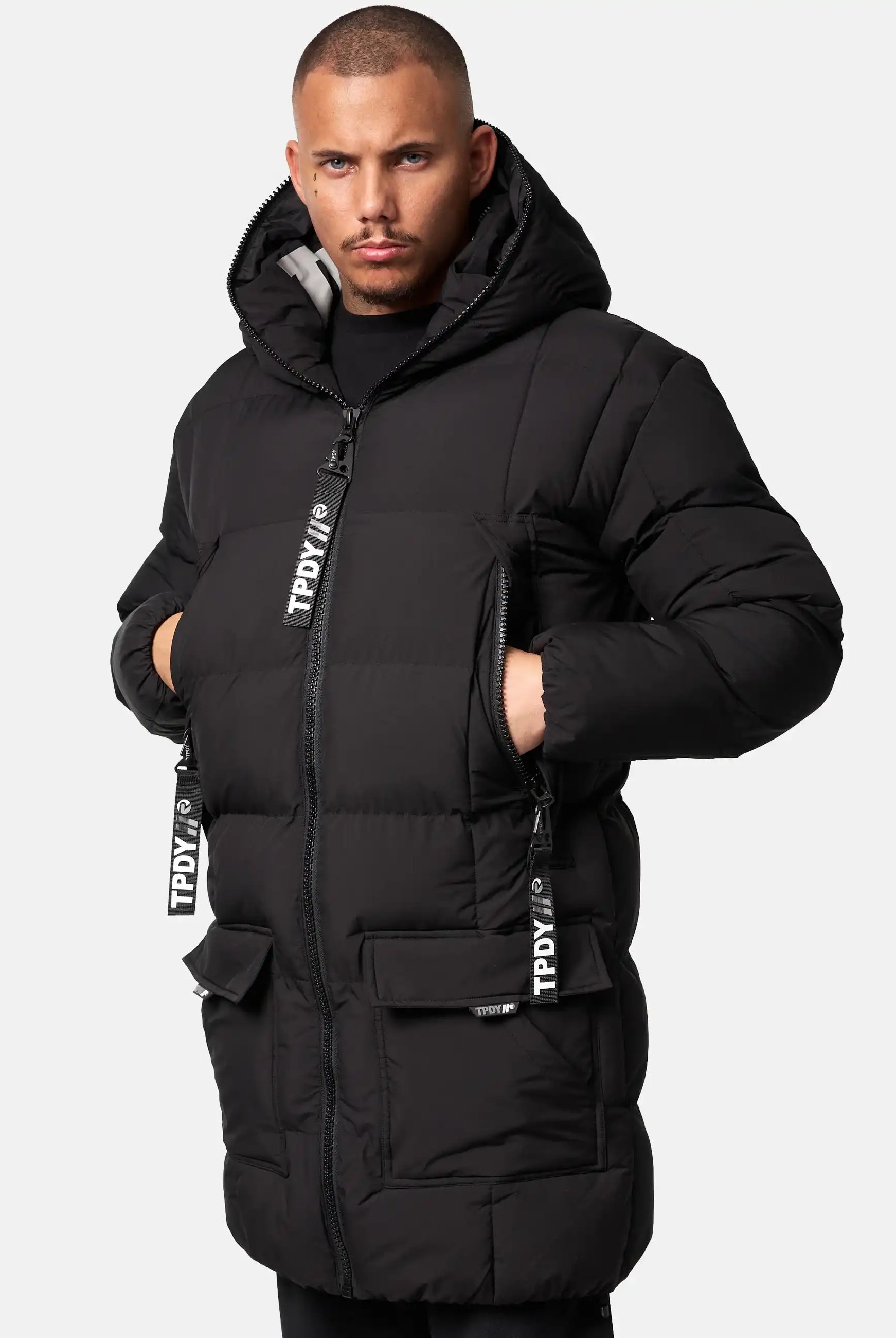 Eine Person, die die trueprodigy Willo Lange Pufferjacke mit Kapuze trägt - schwarz, lang, gesteppt, mit Reißverschluss vorne, großen Taschen und TPDI-gebrandeten Trägern - steht vor einem schlichten hellen Hintergrund.