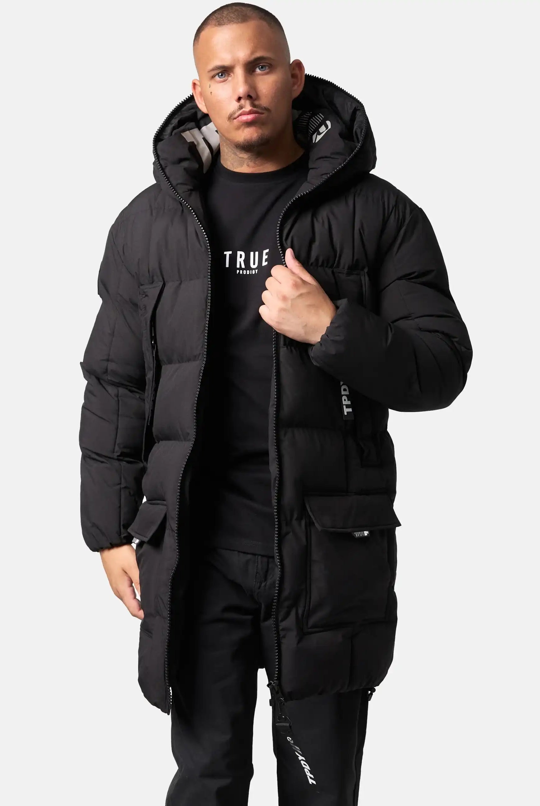 Ein Mann steht vor einem schlichten Hintergrund und trägt die Willo Lange Pufferjacke mit Kapuze von trueprodigy, ein schwarzes T-Shirt und eine schwarze Hose. Er öffnet teilweise den Reißverschluss der Jacke und schaut geradeaus.