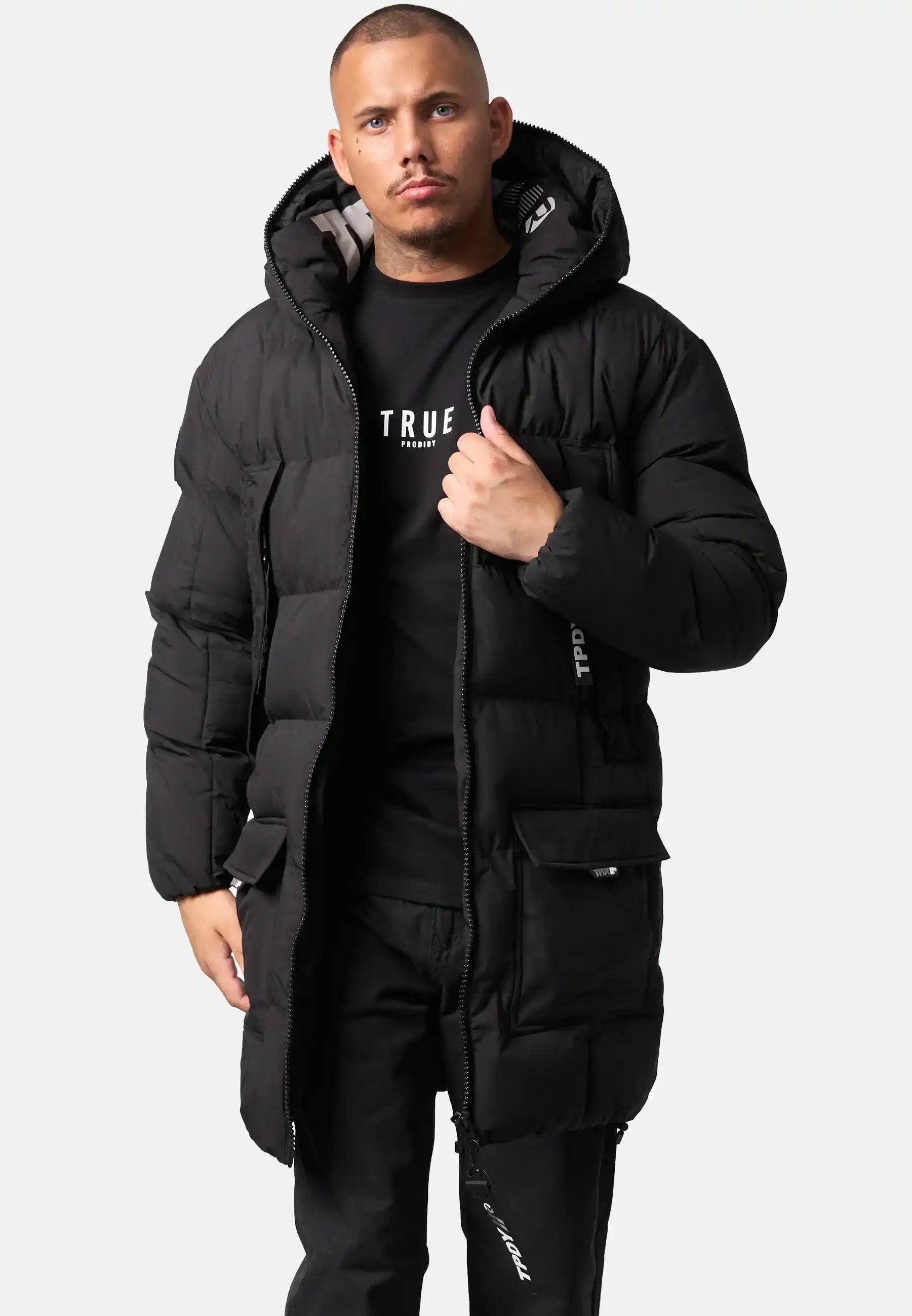 Ein Mann steht vor einem schlichten Hintergrund und trägt die Willo Lange Pufferjacke mit Kapuze von trueprodigy, ein schwarzes T-Shirt und eine schwarze Hose. Er öffnet teilweise den Reißverschluss der Jacke und schaut geradeaus.