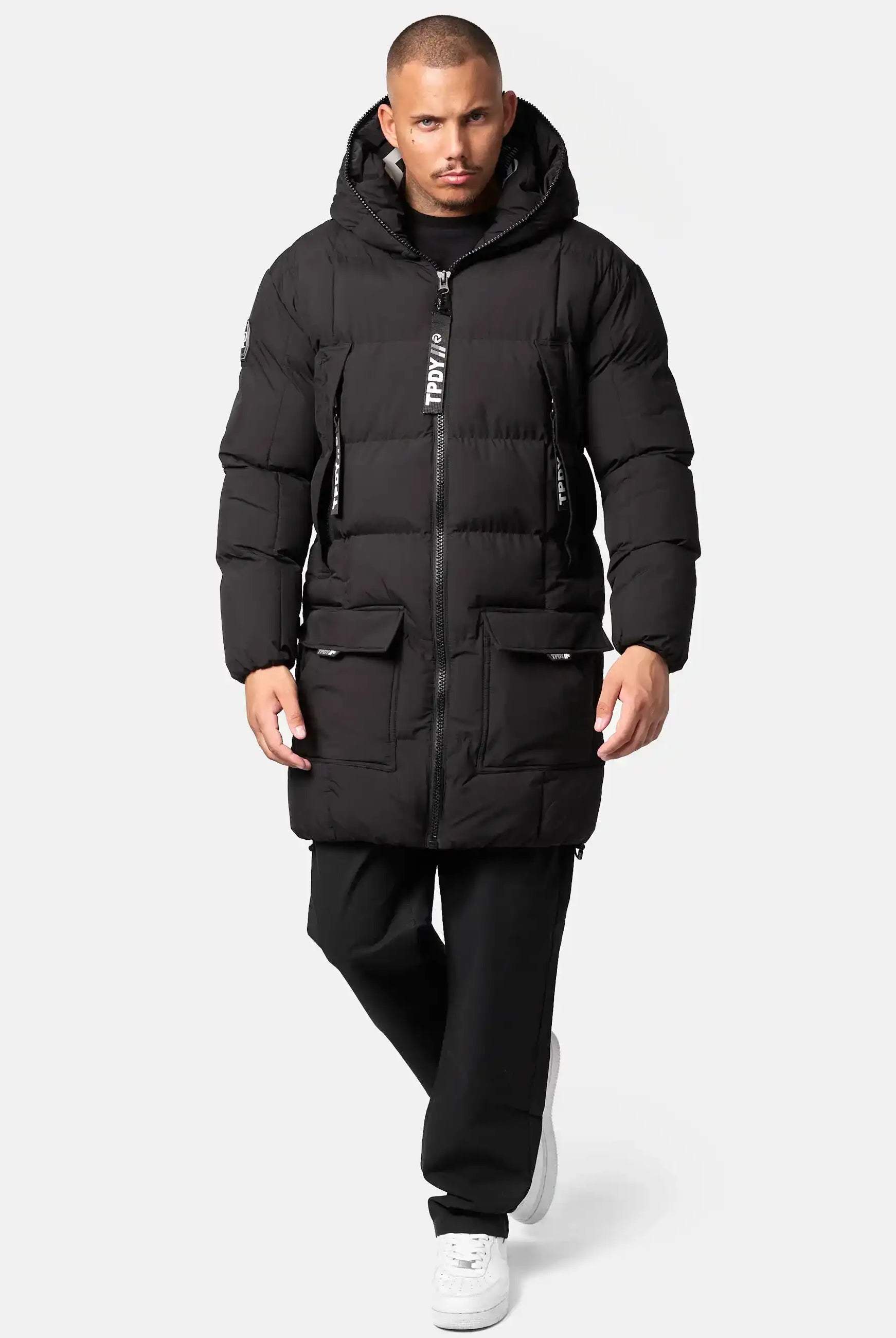 Ein Mann trägt die trueprodigy Willo Lange Pufferjacke mit Kapuze, gepaart mit einer schwarzen Hose und weißen Turnschuhen, vor einem schlichten hellen Hintergrund. Die wasserabweisende Jacke hat einen vertikalen Reißverschluss und große Fronttaschen.