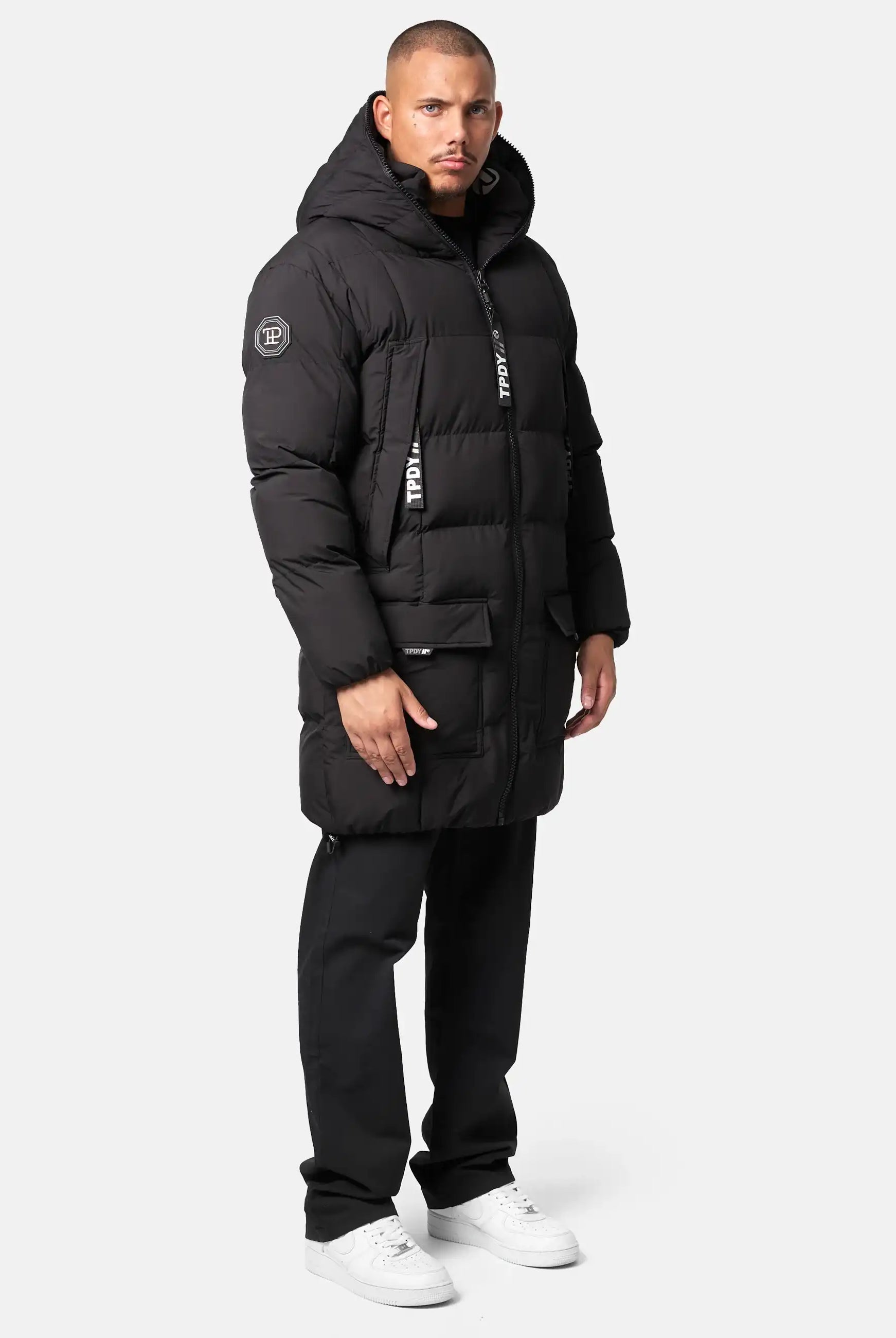 Ein Mann steht mit dem Gesicht nach vorne in einer Trueprodigy Willo Lange Pufferjacke mit Kapuze, einer schwarzen Hose und weißen Turnschuhen vor einem schlichten hellen Hintergrund.