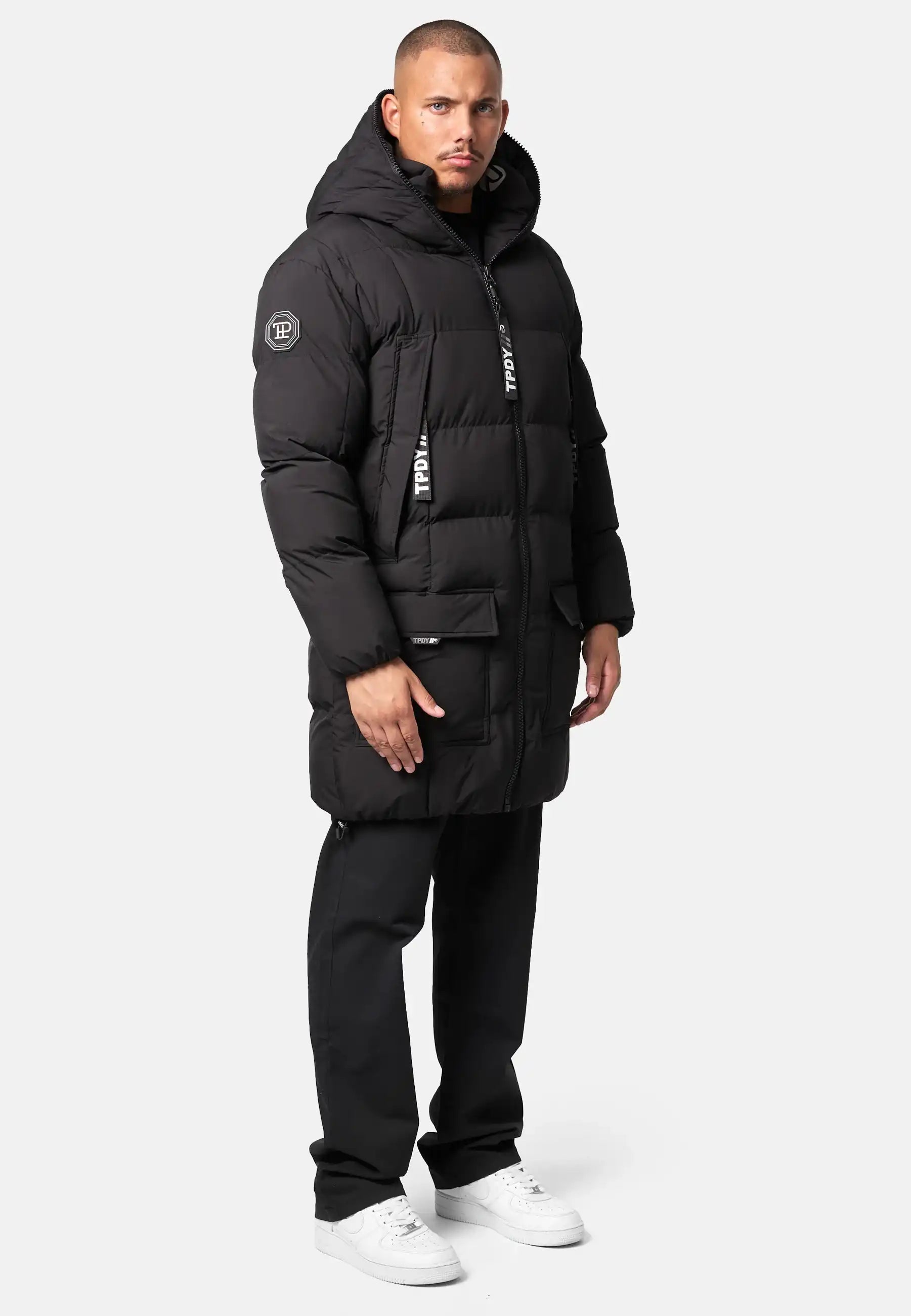 Ein Mann steht mit dem Gesicht nach vorne in einer Trueprodigy Willo Lange Pufferjacke mit Kapuze, einer schwarzen Hose und weißen Turnschuhen vor einem schlichten hellen Hintergrund.