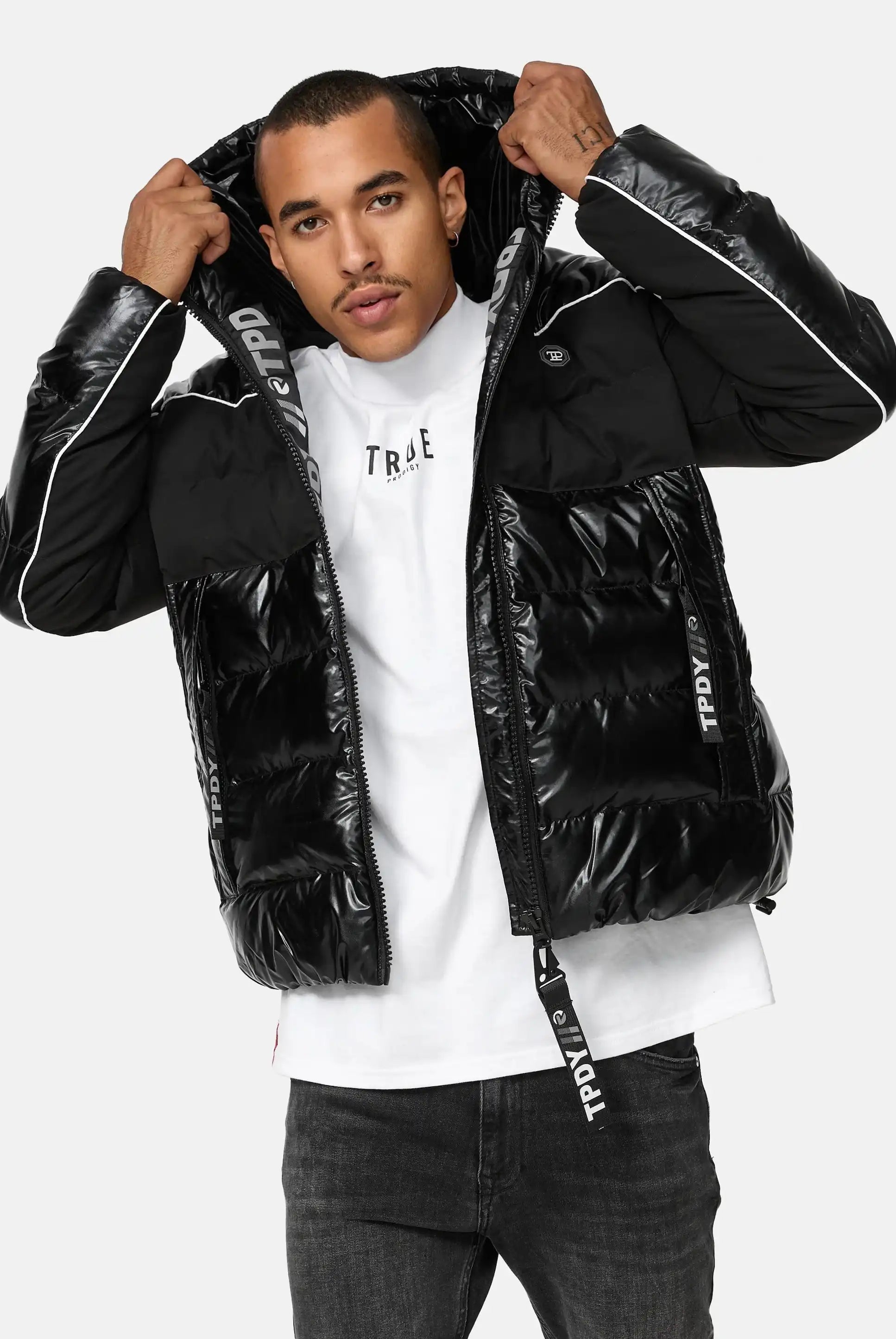 Ein Mann steht mit dem Gesicht nach vorne und trägt eine glänzende schwarze trueprodigy Clark Winterjacke mit Kapuze und Logo mit weißen Paspeln und gebrandeten Reißverschlüssen über einem weißen T-Shirt. Er hält die Kapuze der reflektierenden Jacke mit beiden Händen fest.