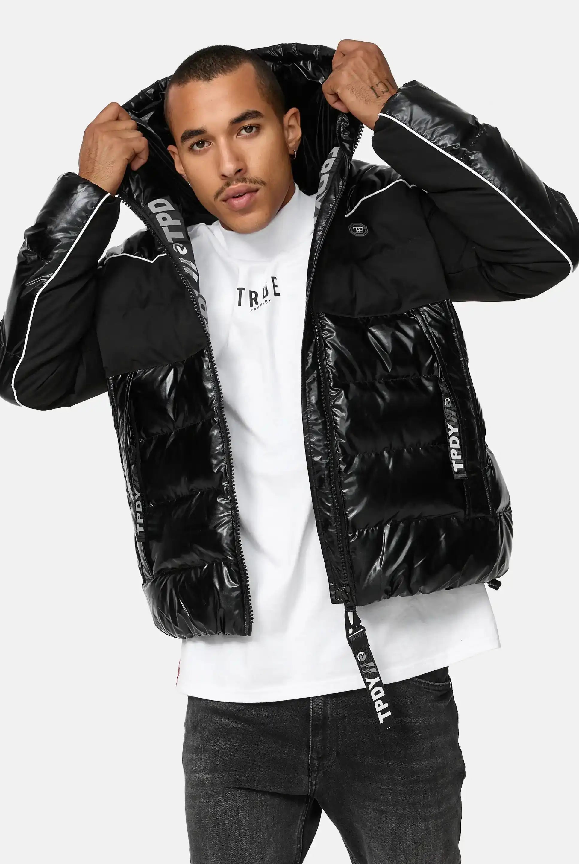 Ein Mann steht mit dem Gesicht nach vorne und trägt eine glänzende schwarze trueprodigy Clark Winterjacke mit Kapuze und Logo mit weißen Paspeln und gebrandeten Reißverschlüssen über einem weißen T-Shirt. Er hält die Kapuze der reflektierenden Jacke mit beiden Händen fest.