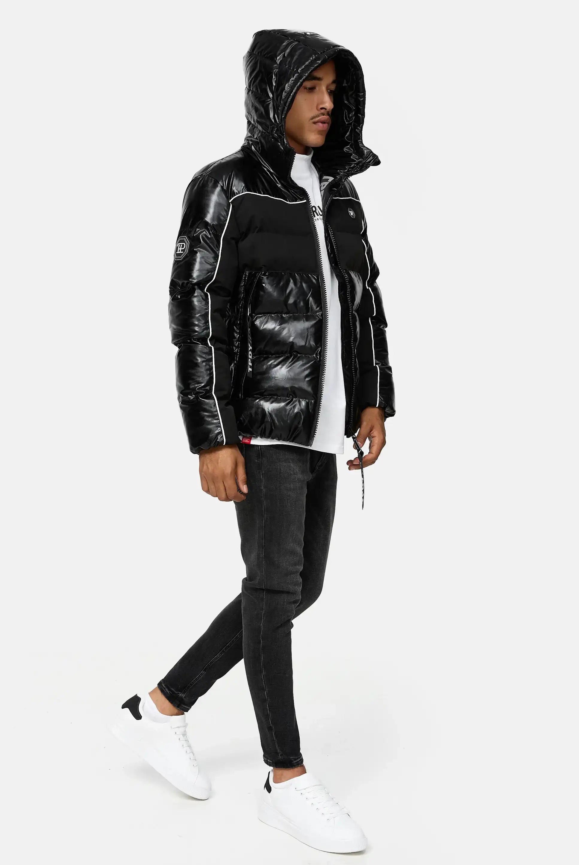 Ein Mann in der trueprodigy Clark Winterjacke mit Kapuze und Logo in glänzendem Schwarz, gepaart mit einem weißen T-Shirt, schwarzen Skinny-Jeans und weißen Turnschuhen, steht vor einem schlichten hellen Hintergrund und schaut nach links, als würde er gehen.