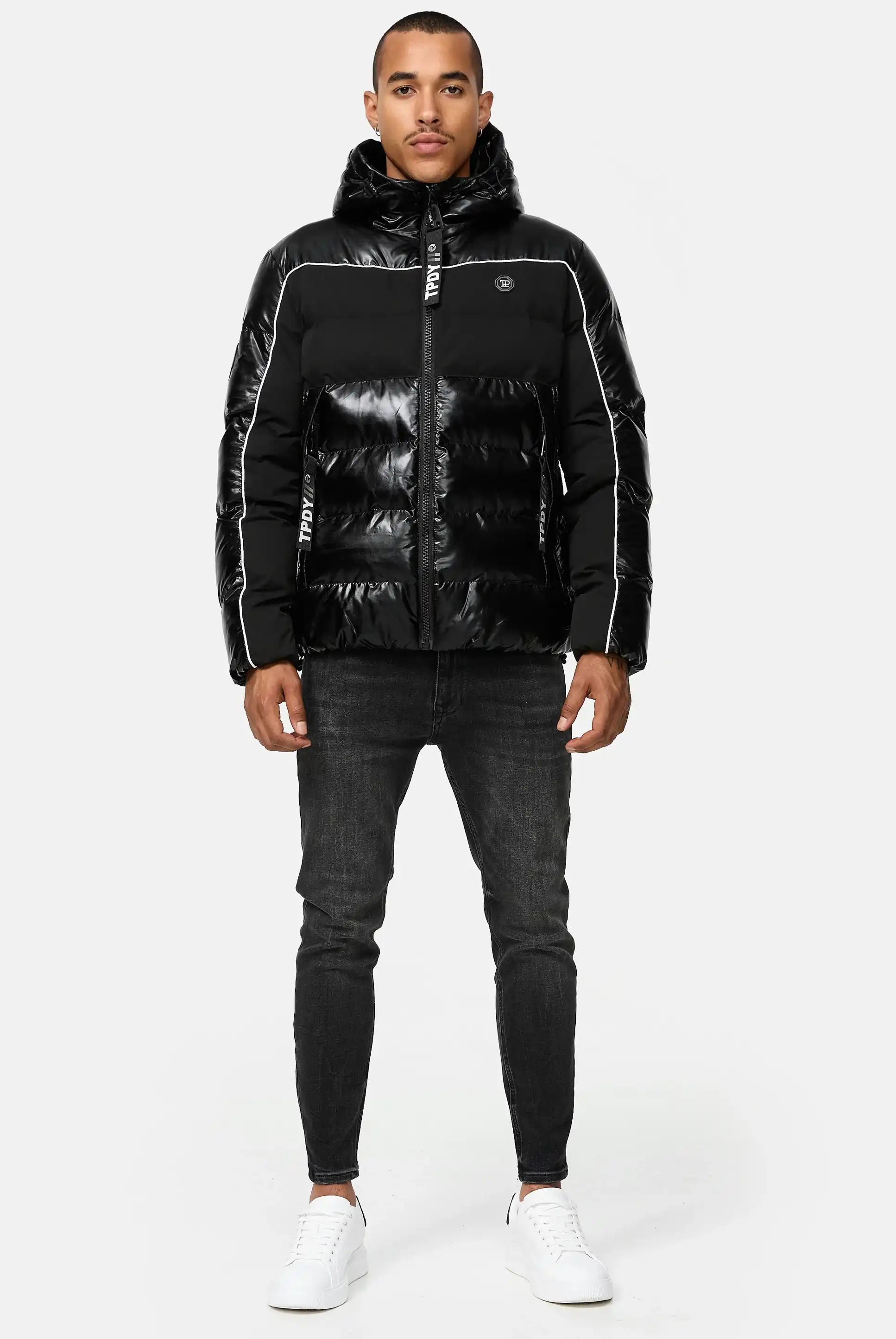 Ein Mann blickt nach vorne und trägt die glänzende schwarze trueprodigy Clark Winterjacke mit Kapuze und Logo mit weißen Paspeln, dunkle Skinny Jeans und weiße Turnschuhe vor einem schlichten hellgrauen Hintergrund.