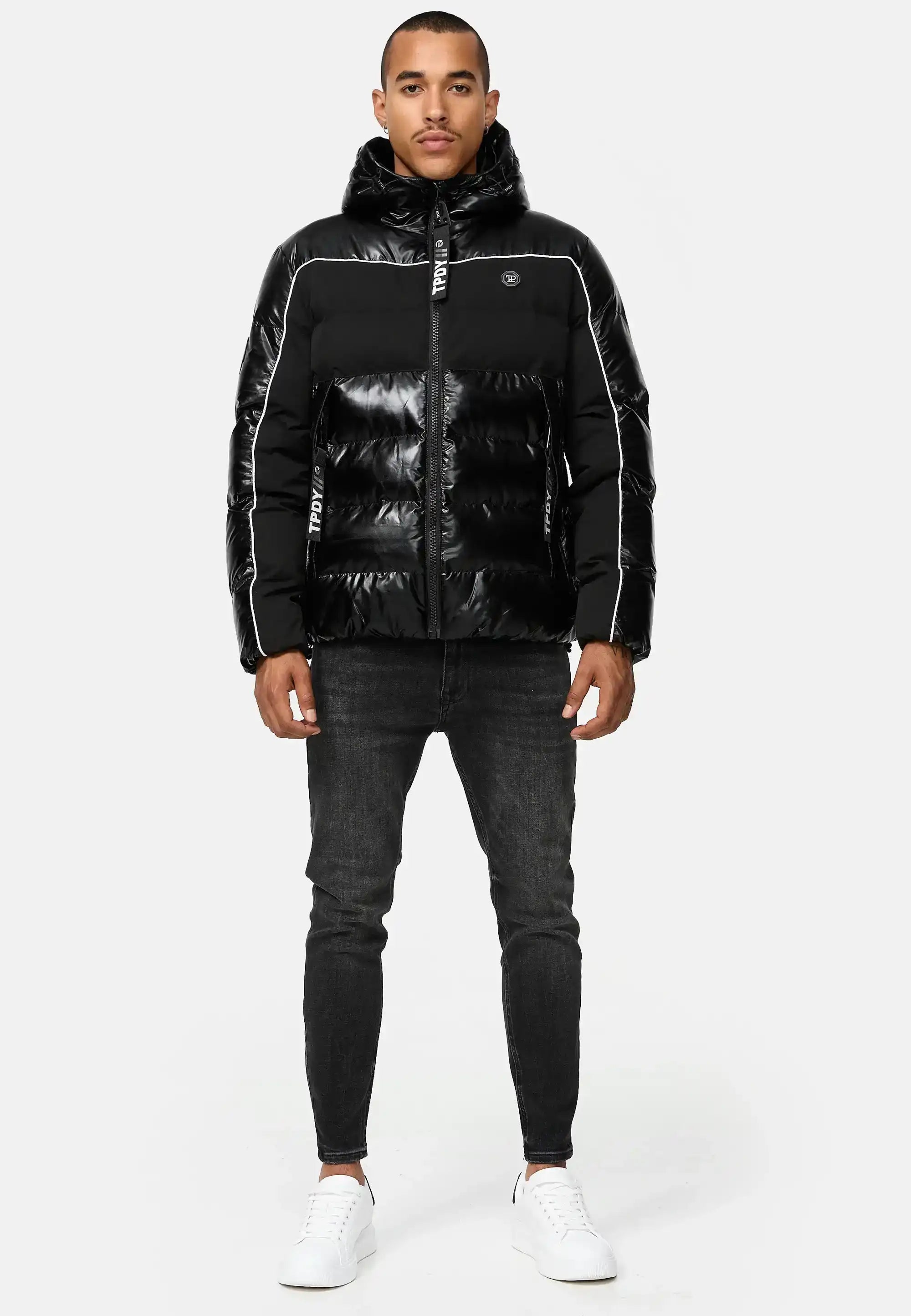 Ein Mann blickt nach vorne und trägt die glänzende schwarze trueprodigy Clark Winterjacke mit Kapuze und Logo mit weißen Paspeln, dunkle Skinny Jeans und weiße Turnschuhe vor einem schlichten hellgrauen Hintergrund.