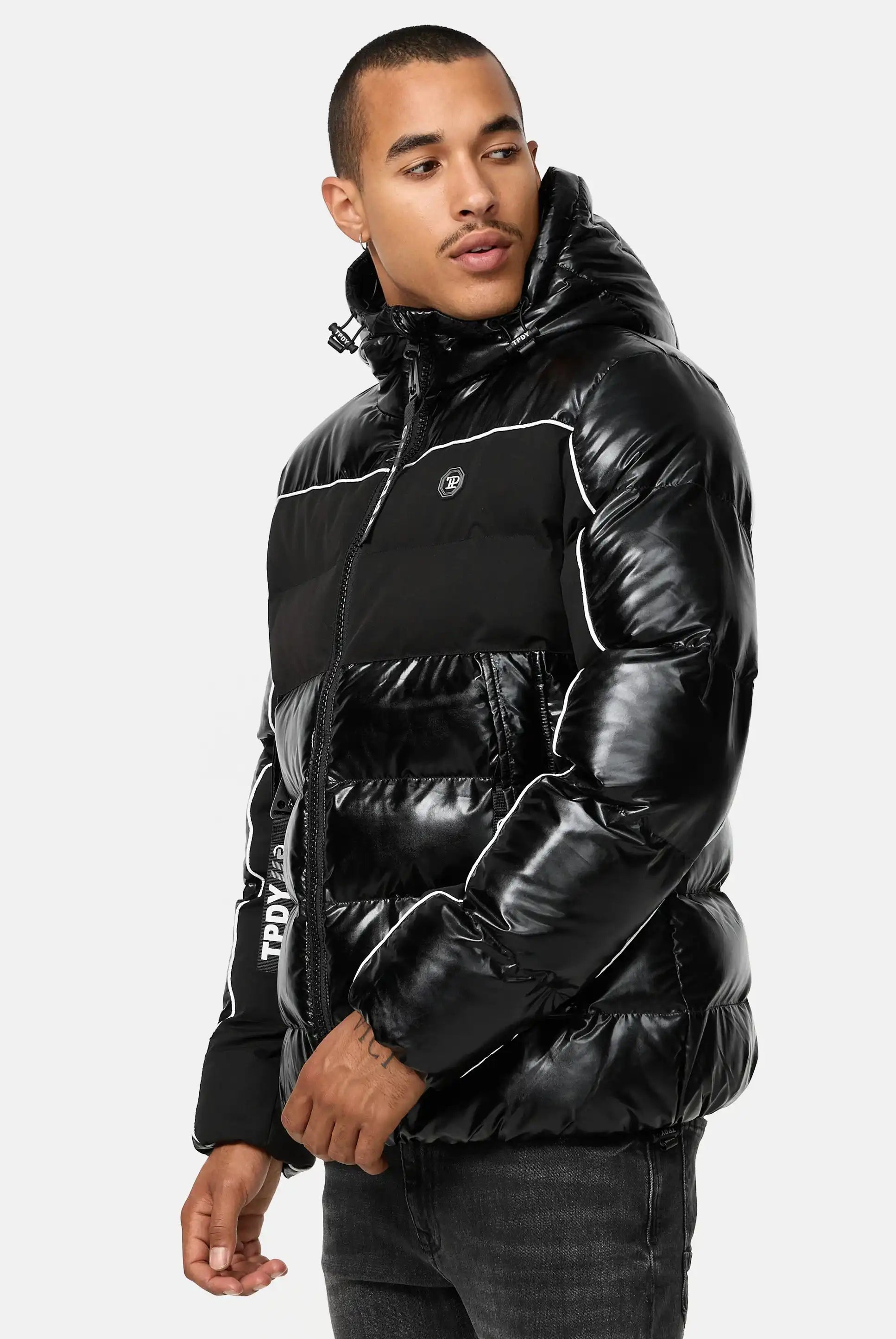 Ein Mann in der Clark Winterjacke mit Kapuze und Logo von trueprodigy - einer glänzenden schwarzen Pufferjacke mit Kapuze und schwarzen Jeans - steht mit neutralem Gesichtsausdruck vor einem schlichten weißen Hintergrund.