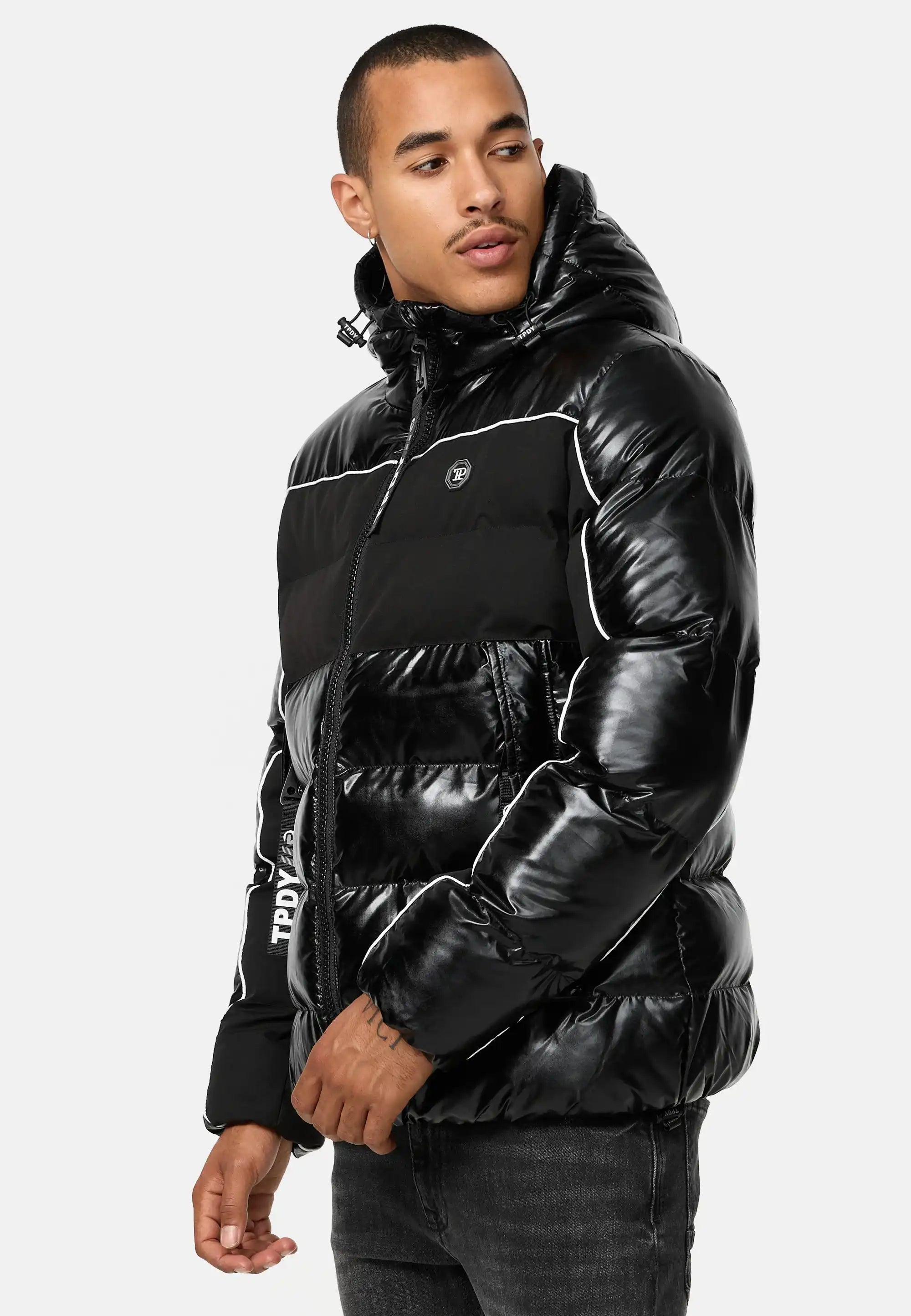 Ein Mann in der Clark Winterjacke mit Kapuze und Logo von trueprodigy - einer glänzenden schwarzen Pufferjacke mit Kapuze und schwarzen Jeans - steht mit neutralem Gesichtsausdruck vor einem schlichten weißen Hintergrund.