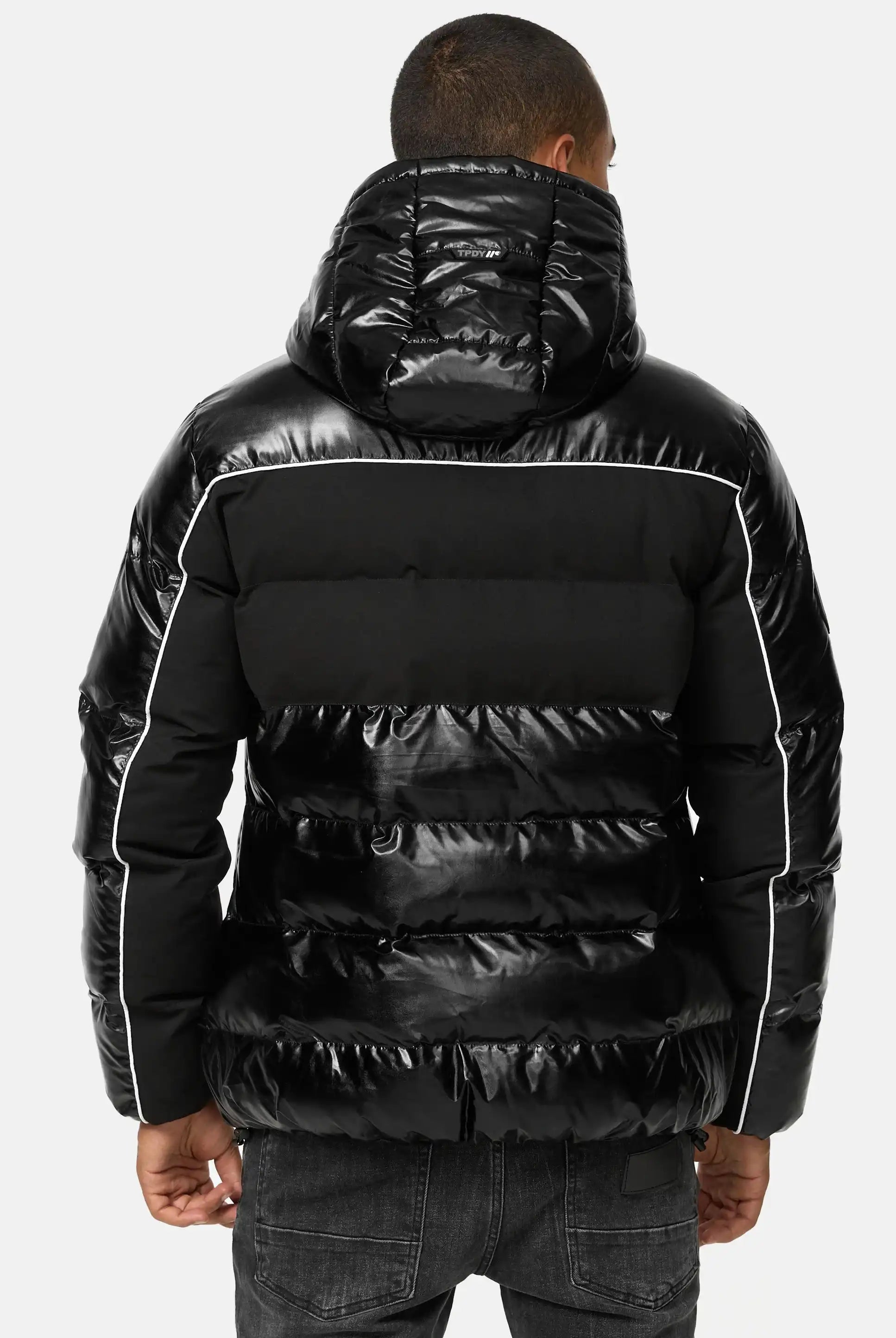 Man sieht eine Person von hinten, die die trueprodigy Clark Winterjacke mit Kapuze und Logo trägt, eine glänzende schwarze Jacke mit weißen Paspelierungen. Diese gesteppte Winterjacke lässt sich stilvoll mit dunklen Jeans kombinieren und sorgt für einen eleganten Winterlook.