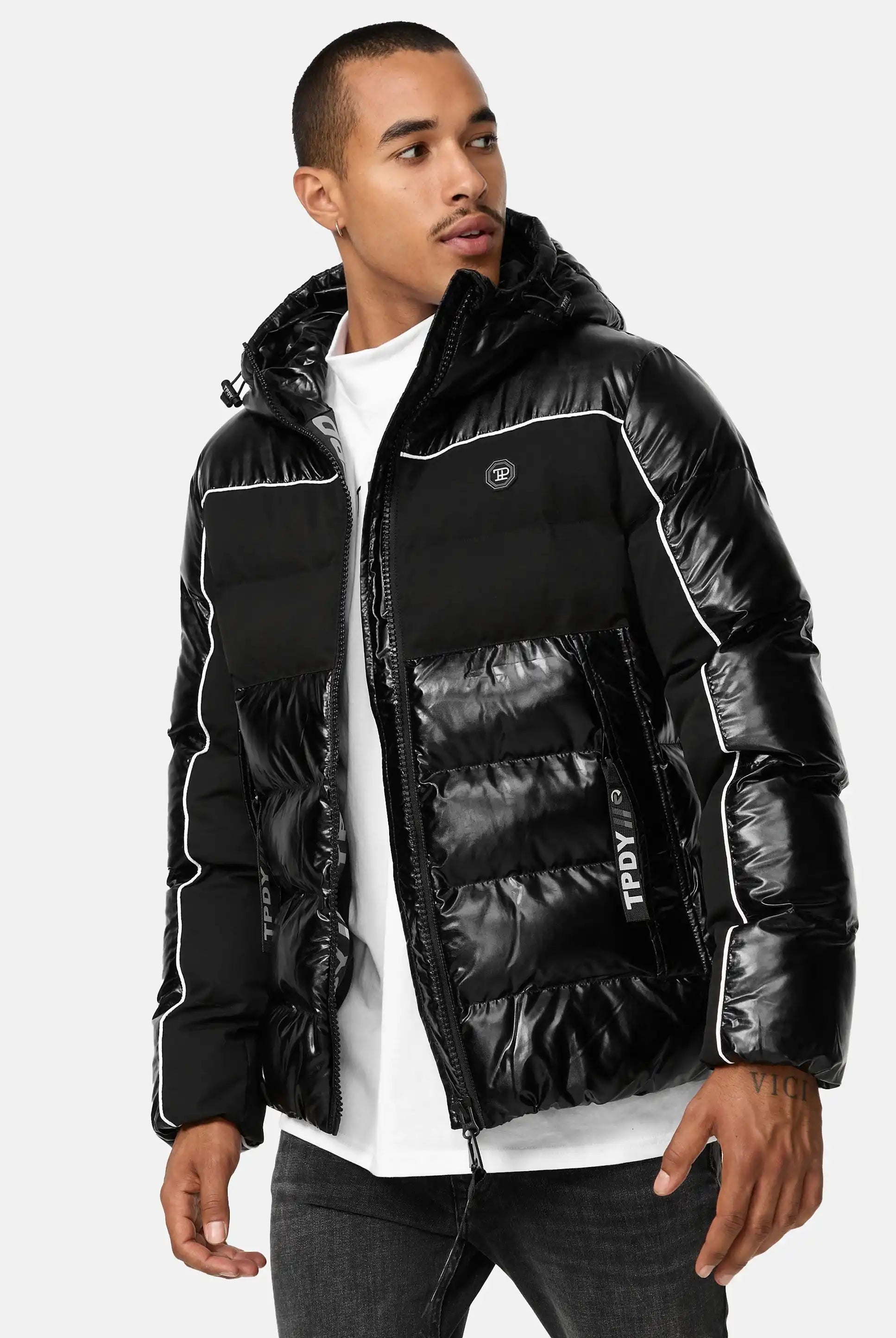 Ein Mann in der glänzenden Clark Winterjacke mit Kapuze und Logo von trueprodigy mit weißen Paspeln und einer Kapuze über einem weißen Hemd und einer dunklen Jeans steht leicht seitlich vor einem unifarbenen Hintergrund.