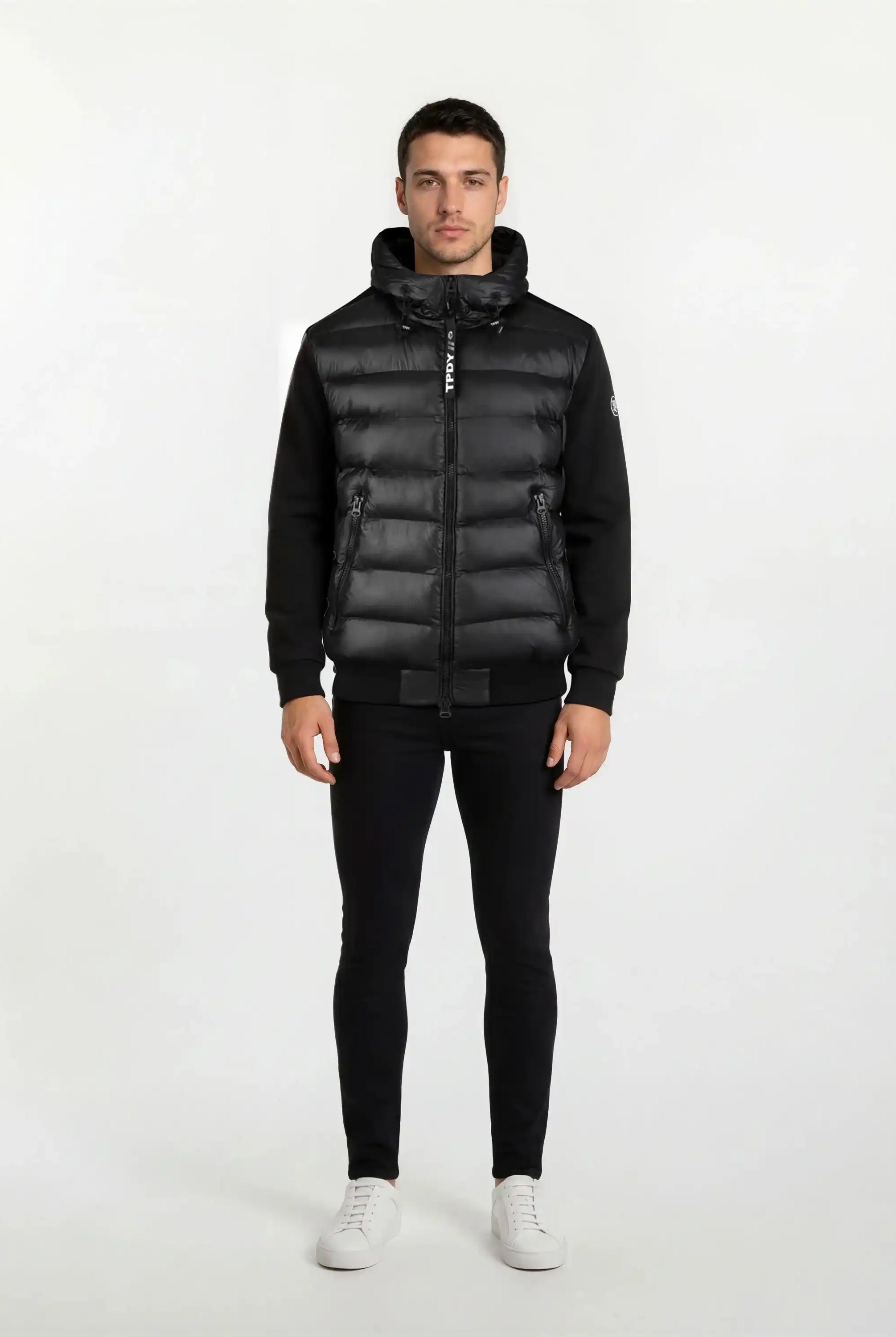 A man steht frontal vor weißem Hintergrund, trägt eine gesteppte Herren Übergangsjacke für urbanen City-Style von trueprodigy mit Kapuze, dazu schwarze Hosen und weiße Sneaker. Seine Arme hängen locker an den Seiten.