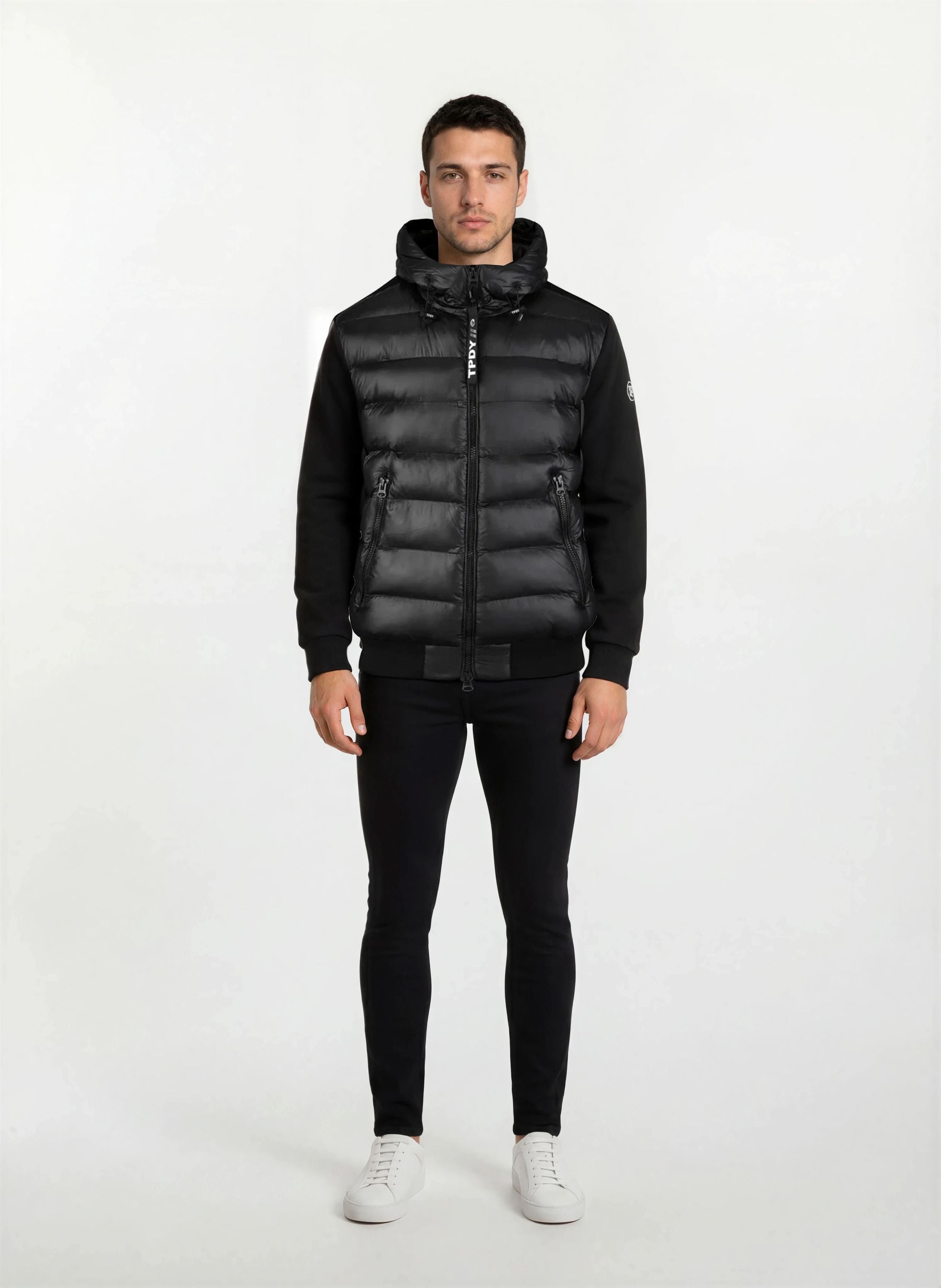 A man steht frontal vor weißem Hintergrund, trägt eine gesteppte Herren Übergangsjacke für urbanen City-Style von trueprodigy mit Kapuze, dazu schwarze Hosen und weiße Sneaker. Seine Arme hängen locker an den Seiten.