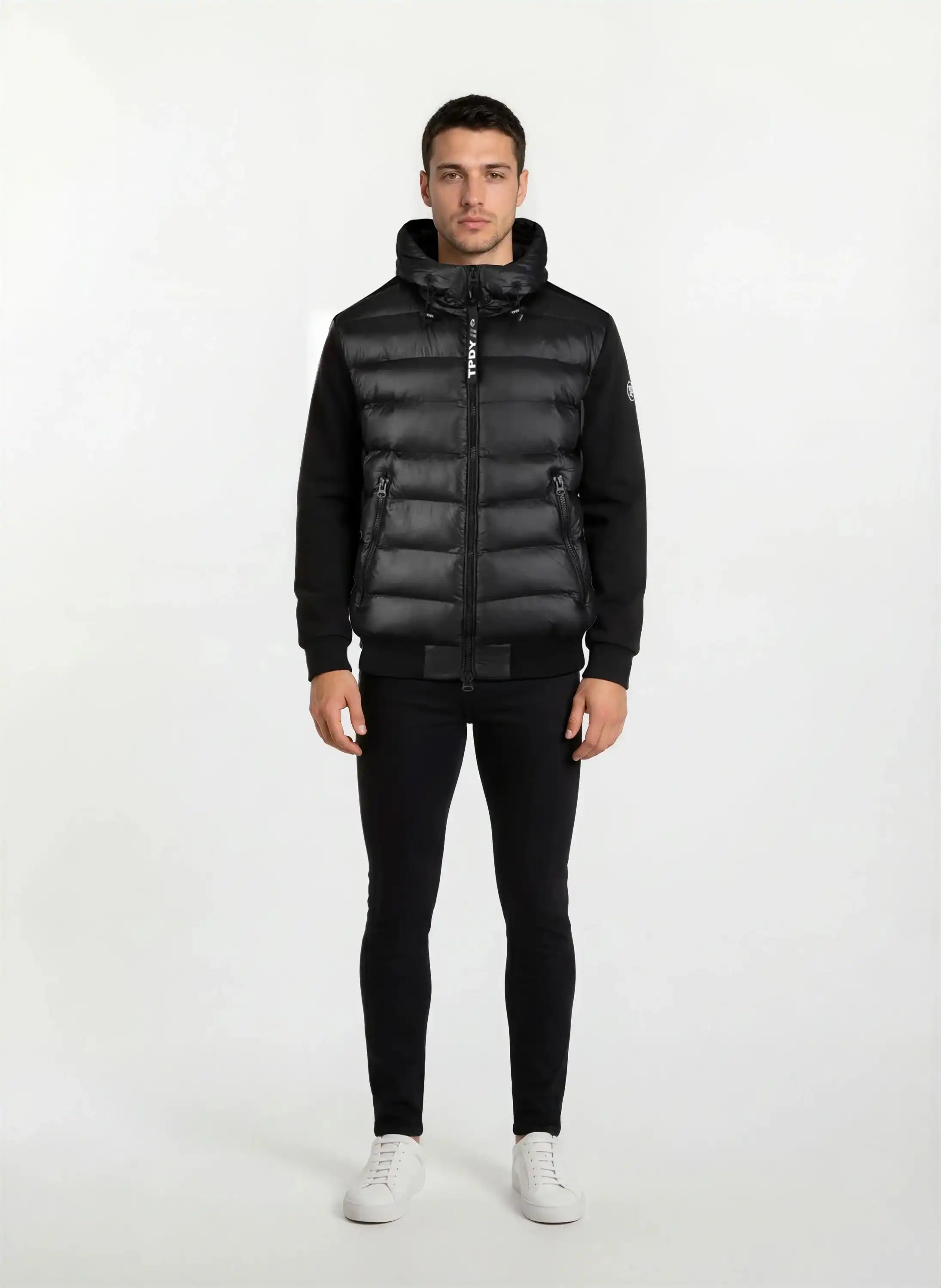 A man steht frontal vor weißem Hintergrund, trägt eine gesteppte Herren Übergangsjacke für urbanen City-Style von trueprodigy mit Kapuze, dazu schwarze Hosen und weiße Sneaker. Seine Arme hängen locker an den Seiten.