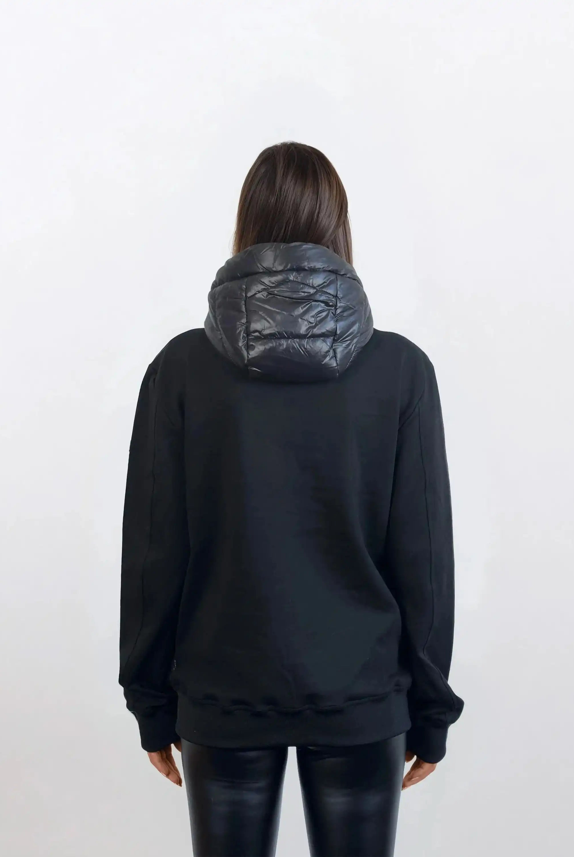 Eine Person mit langen braunen Haaren, von hinten gesehen, trägt eine trueprodigy Oversized Damen Bomber Jacke im urbanen Materialmix-Design und schwarze Kunstlederleggings vor einem schlichten hellen Hintergrund.