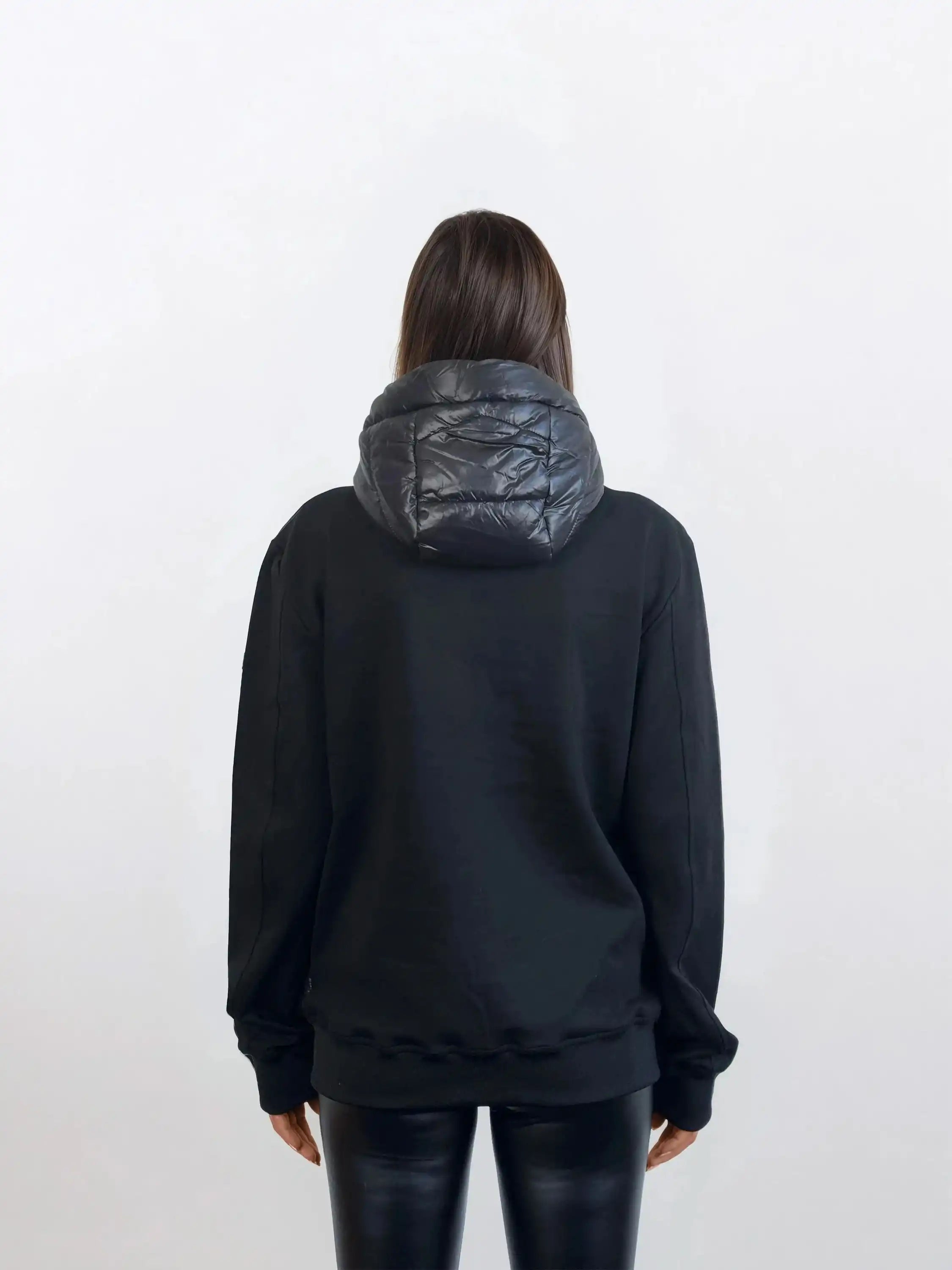 Eine Person mit langen braunen Haaren, von hinten gesehen, trägt eine trueprodigy Oversized Damen Bomber Jacke im urbanen Materialmix-Design und schwarze Kunstlederleggings vor einem schlichten hellen Hintergrund.