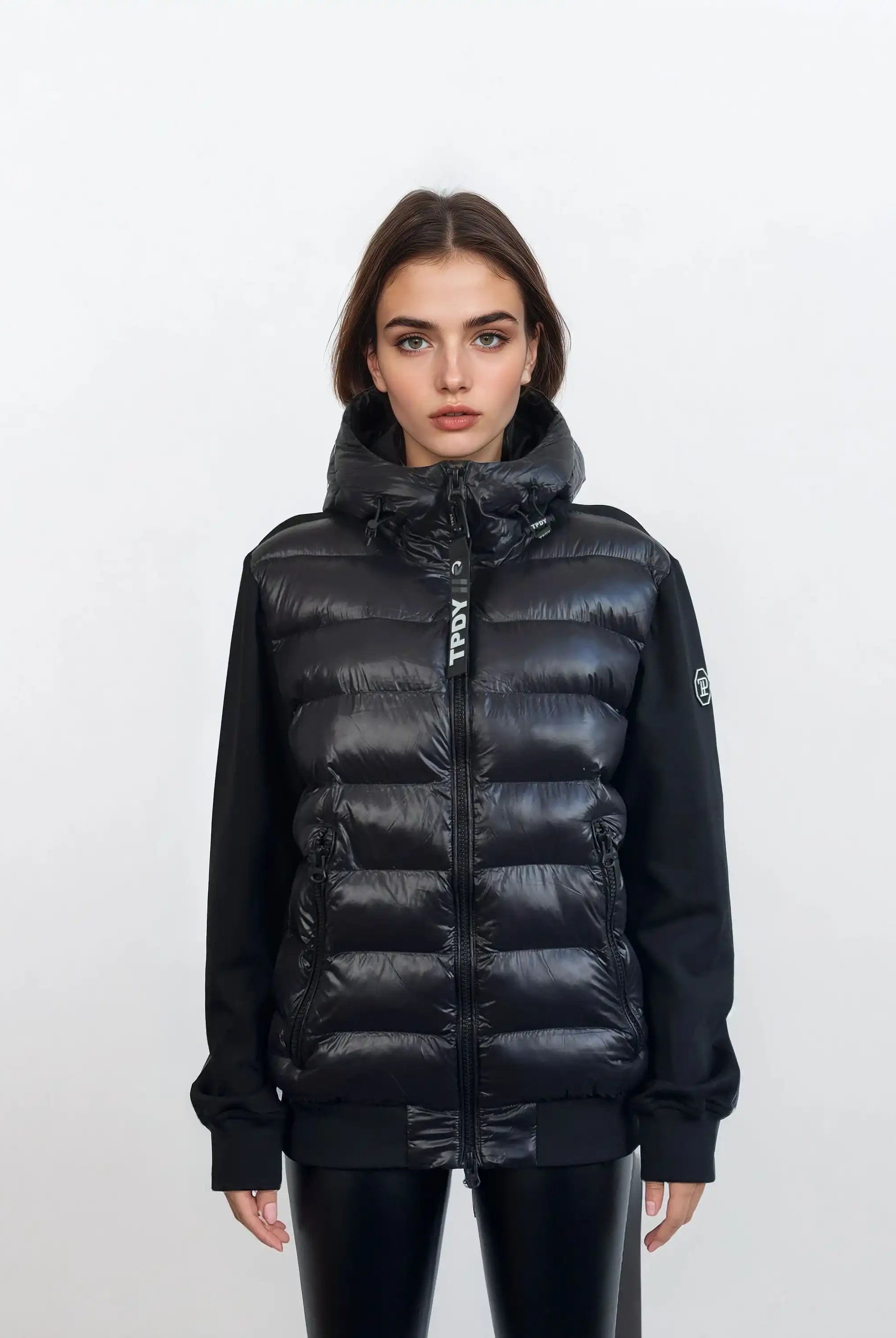 Eine Frau mit glattem braunem Haar steht vor einem weißen Hintergrund, trägt die trueprodigy Oversized Damen Bomber Jacke im urbanen Materialmix-Design mit schwarzen Ärmeln und schwarzer Hose und schaut direkt in die Kamera.