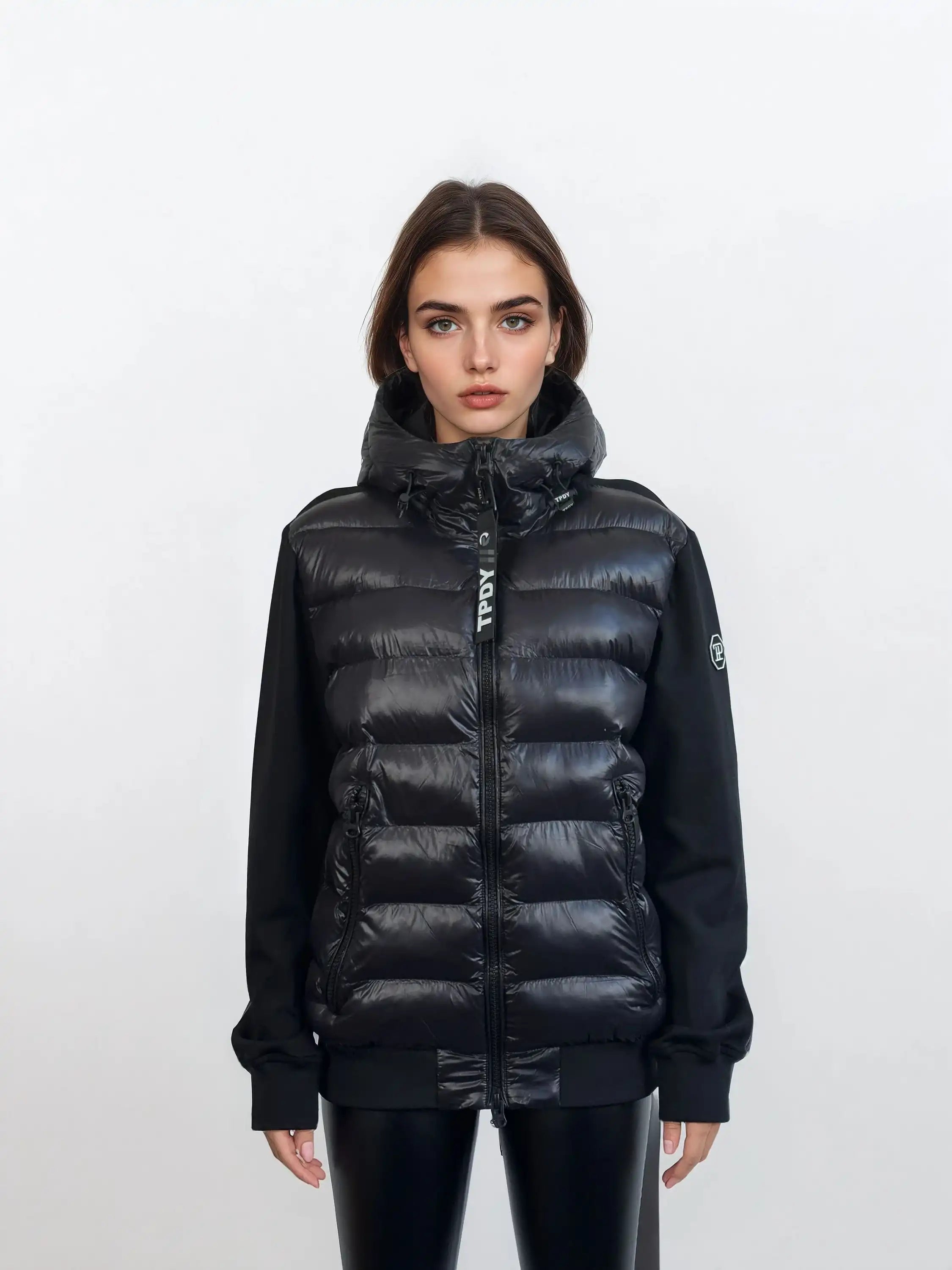 Eine Frau mit glattem braunem Haar steht vor einem weißen Hintergrund, trägt die trueprodigy Oversized Damen Bomber Jacke im urbanen Materialmix-Design mit schwarzen Ärmeln und schwarzer Hose und schaut direkt in die Kamera.