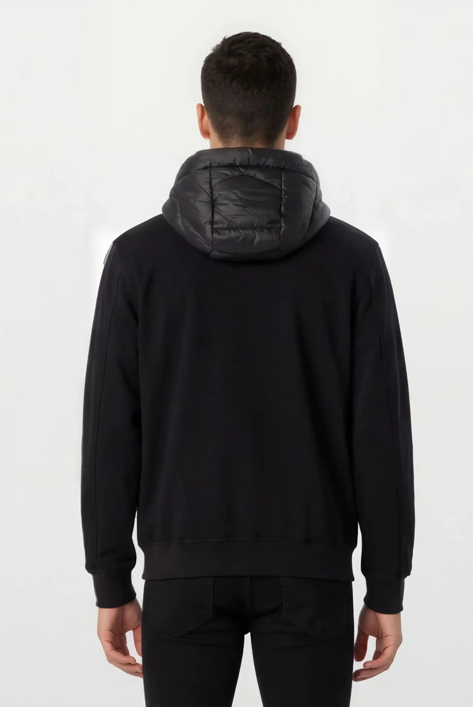 Eine Person steht mit dem Rücken zur Kamera und trägt eine gesteppte trueprodigy Herren Übergangsjacke für urbanen City-Style sowie schwarze Hosen vor schlicht weißem Hintergrund.