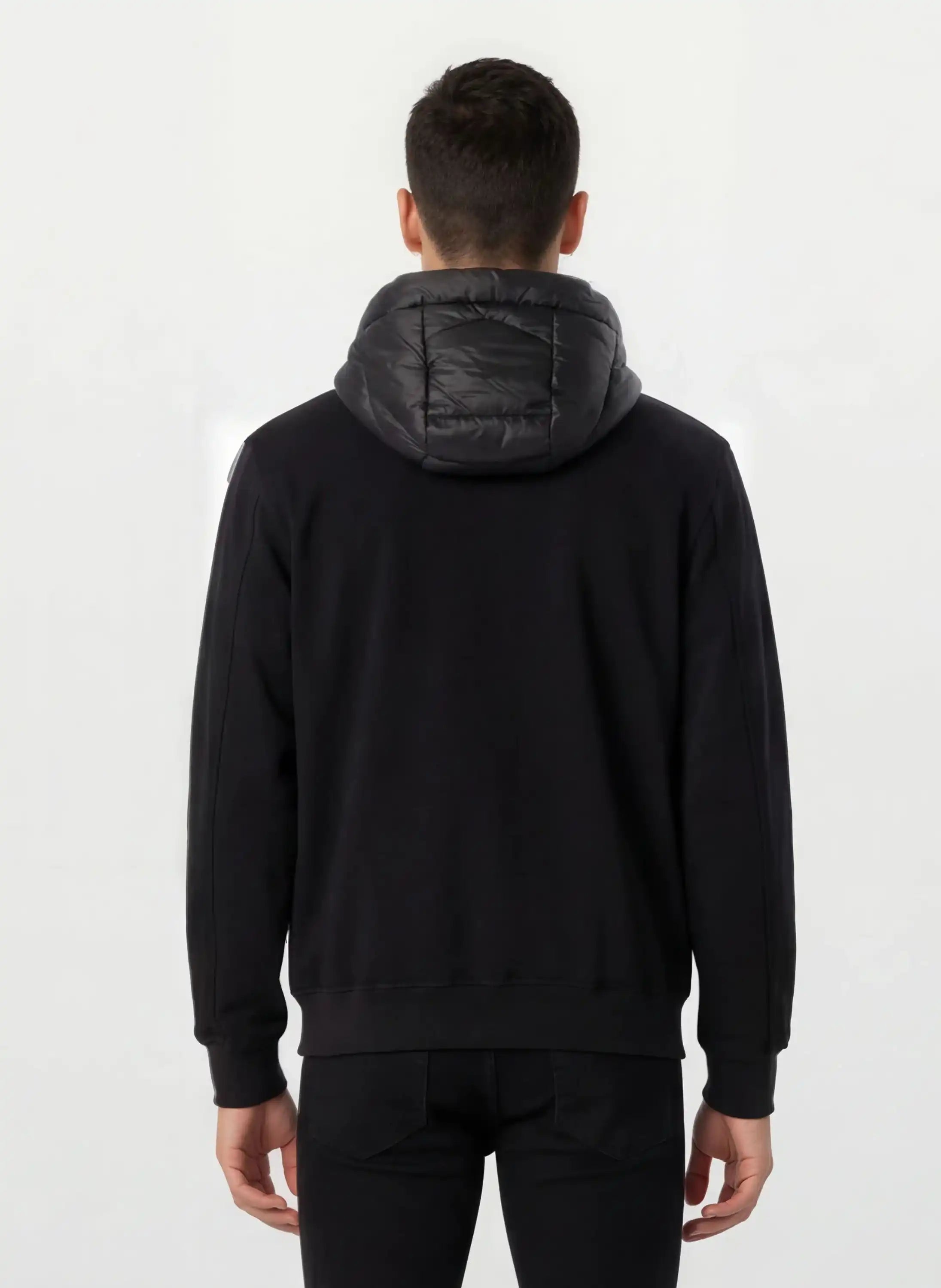 Eine Person steht mit dem Rücken zur Kamera und trägt eine gesteppte trueprodigy Herren Übergangsjacke für urbanen City-Style sowie schwarze Hosen vor schlicht weißem Hintergrund.