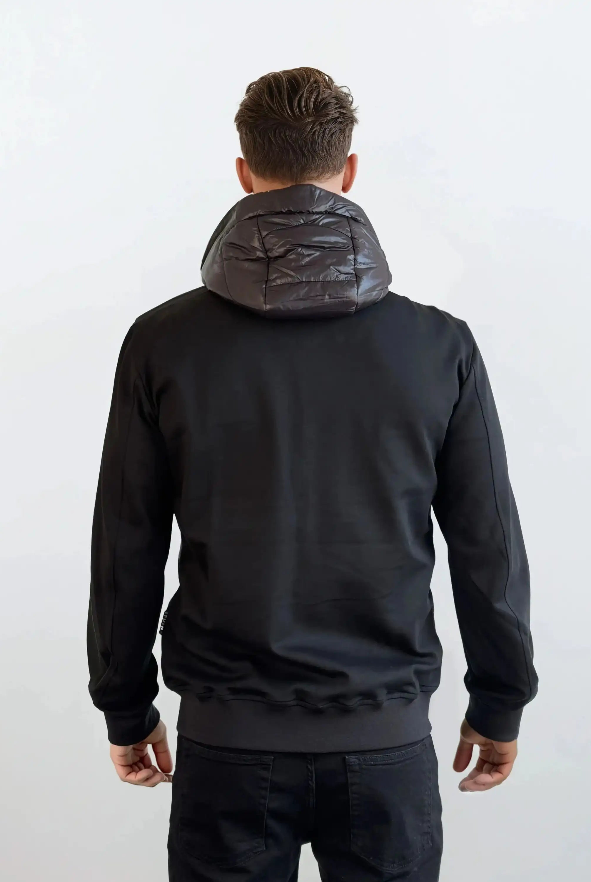 Ein Mann steht mit dem Rücken zur Kamera, bekleidet mit einer trueprodigy Gesteppte Herren Übergangsjacke für urbanen City-Style und einer schwarzen Hose. Der Hintergrund ist einfarbig weiß.