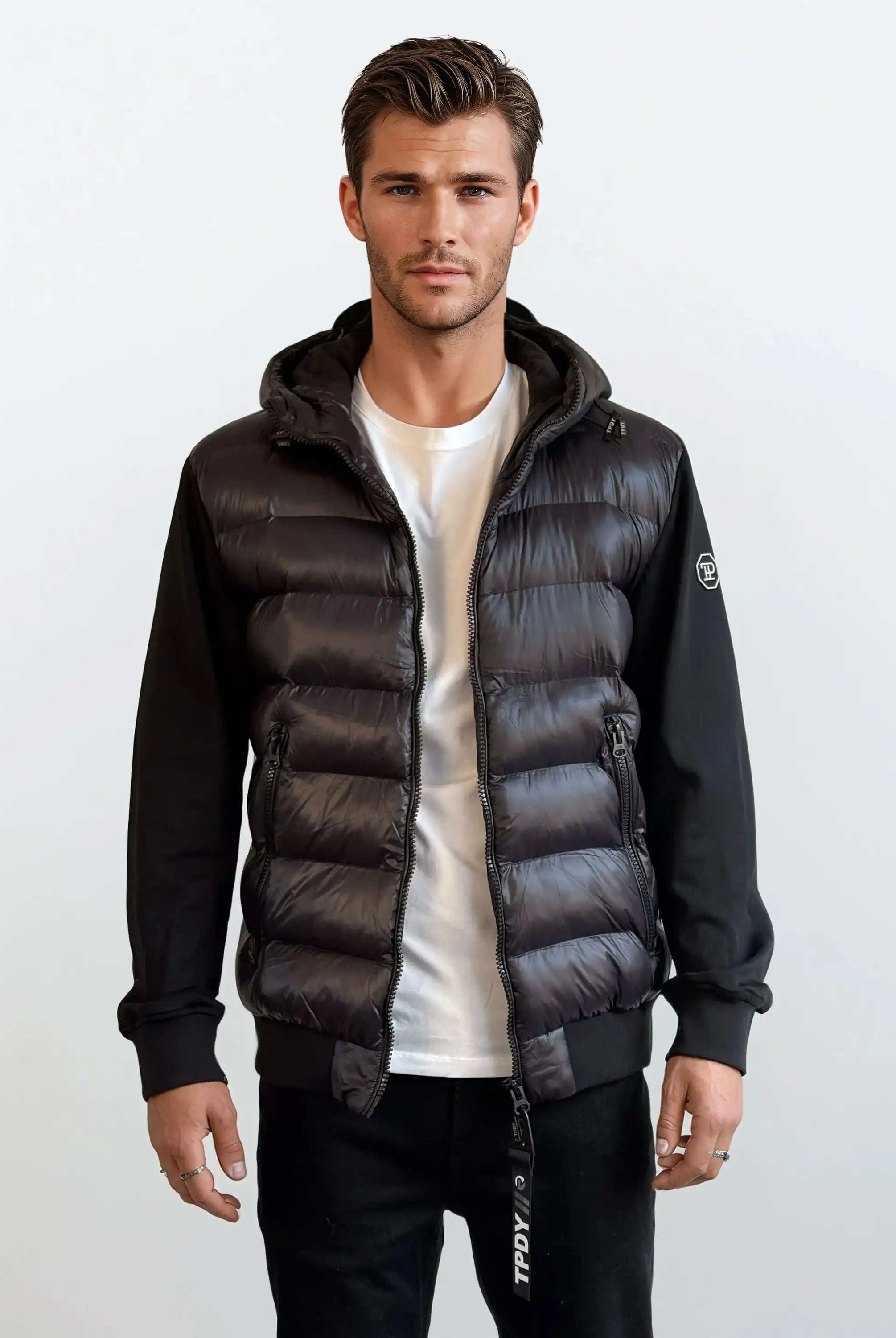 Ein Mann trägt eine schwarze Gesteppte Herren Übergangsjacke für urbanen City-Style von trueprodigy mit weißem T-Shirt und schwarzen Hosen. Der Hintergrund ist schlicht und hell gehalten.