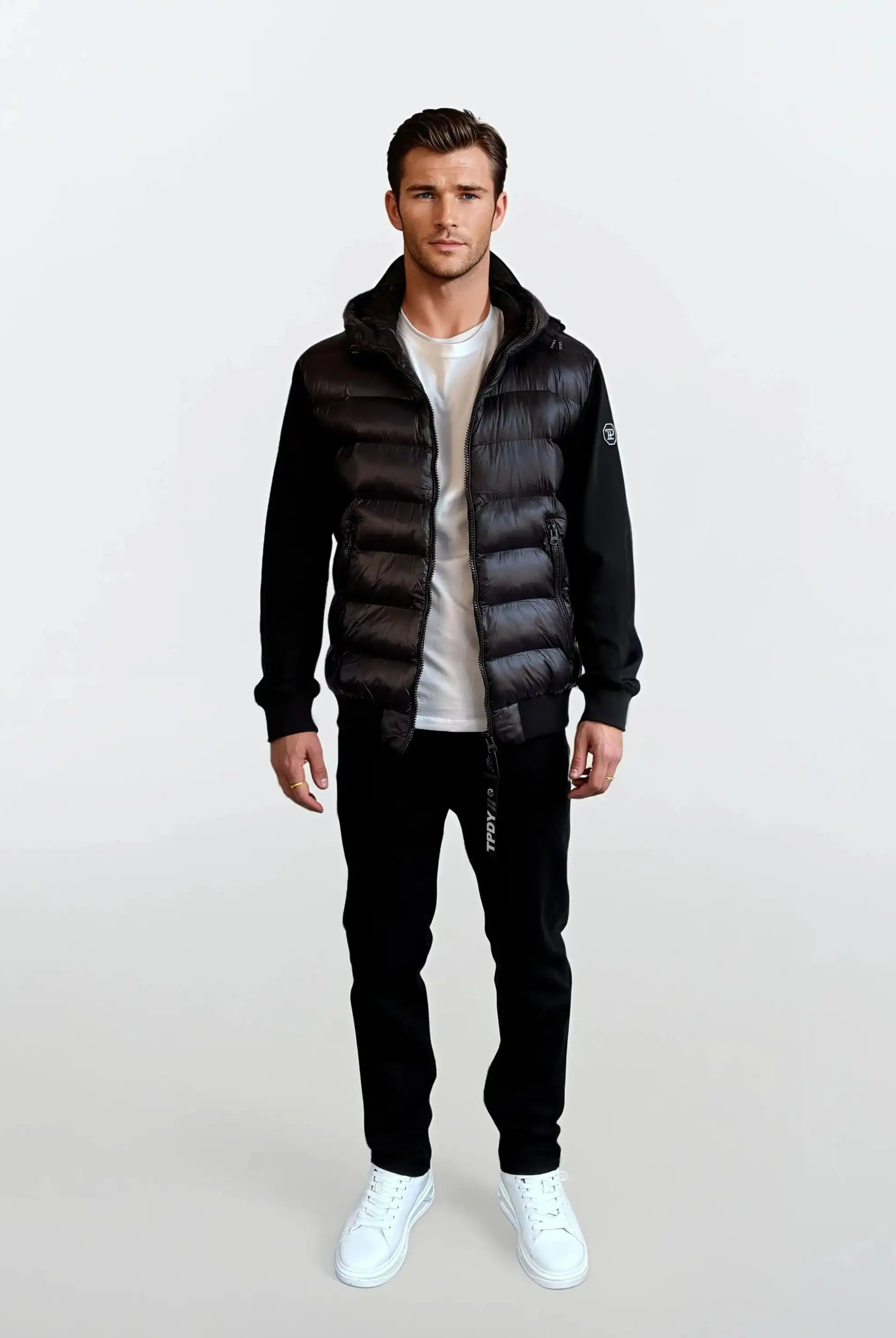 Ein Mann blickt nach vorne in einer trueprodigy Gesteppte Herren Übergangsjacke für urbanen City-Style über einem weißen T-Shirt, einer schwarzen Hose und weißen Sneakers. Der Hintergrund ist schlicht und hell.