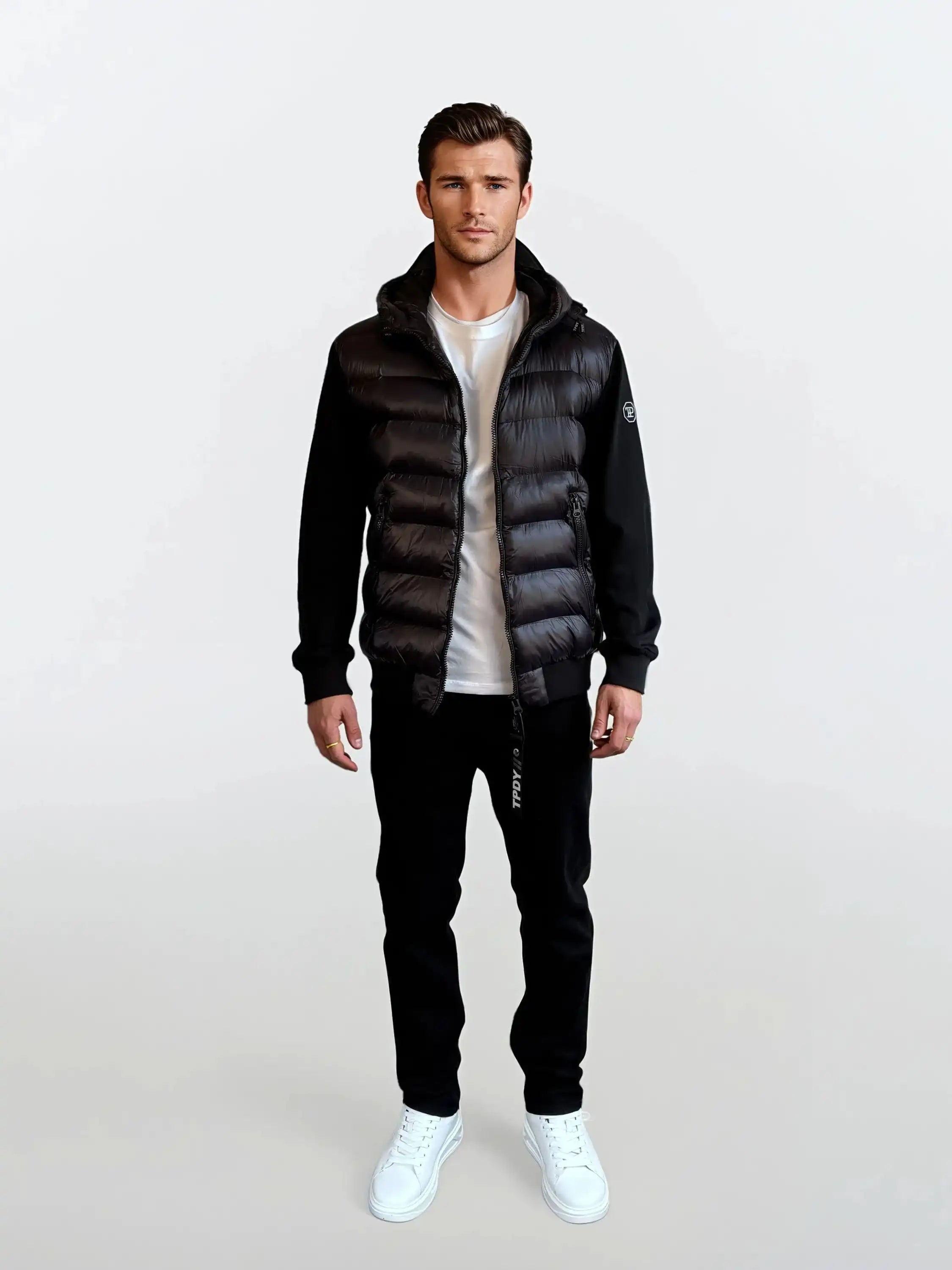 Ein Mann blickt nach vorne in einer trueprodigy Gesteppte Herren Übergangsjacke für urbanen City-Style über einem weißen T-Shirt, einer schwarzen Hose und weißen Sneakers. Der Hintergrund ist schlicht und hell.