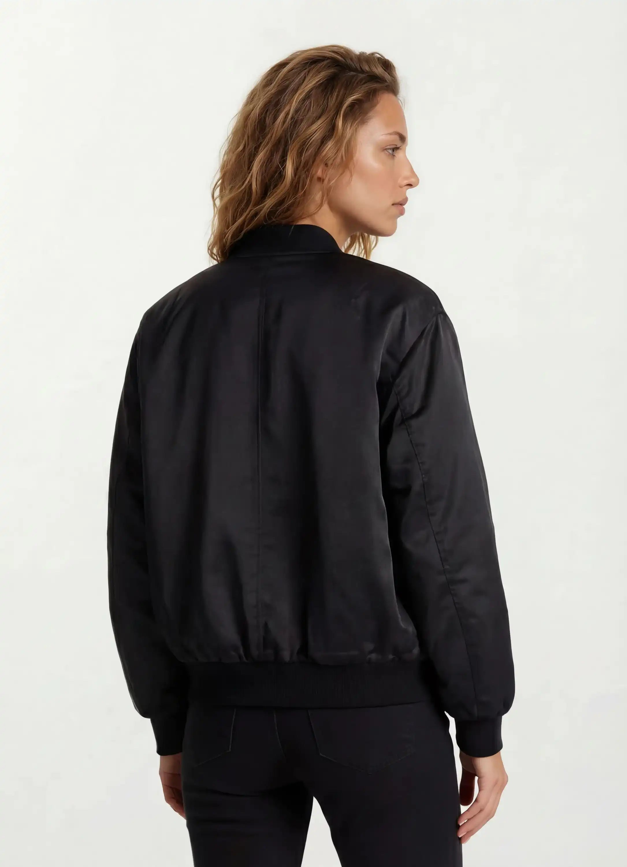 Eine Person mit gewelltem, schulterlangem Haar wird von hinten gezeigt, die eine Elegante Damen Bomber Jacke mit glänzender Oberfläche von trueprodigy vor einem schlichten, hellen Hintergrund trägt.