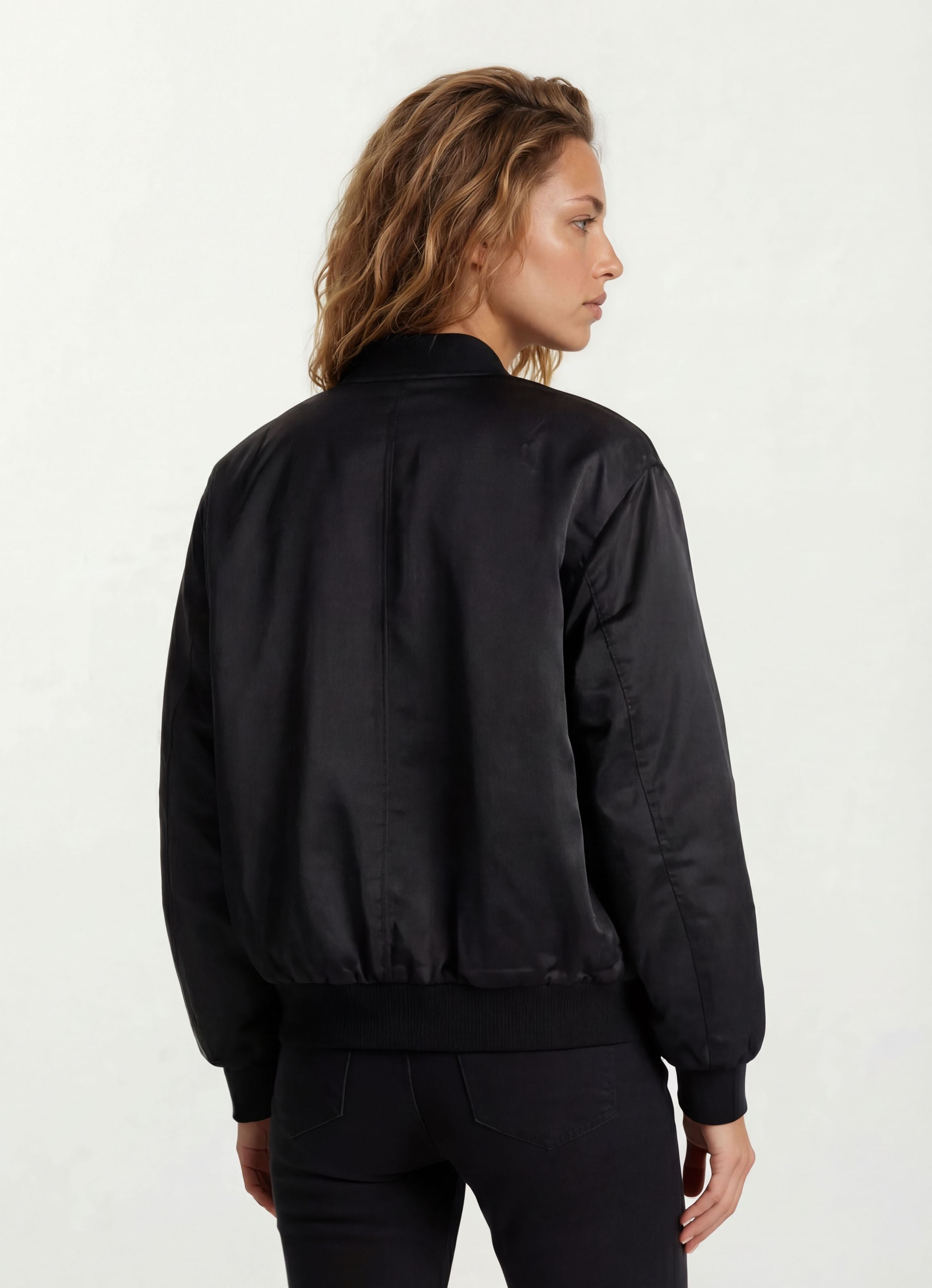 Eine Person mit gewelltem, schulterlangem Haar wird von hinten gezeigt, die eine Elegante Damen Bomber Jacke mit glänzender Oberfläche von trueprodigy vor einem schlichten, hellen Hintergrund trägt.